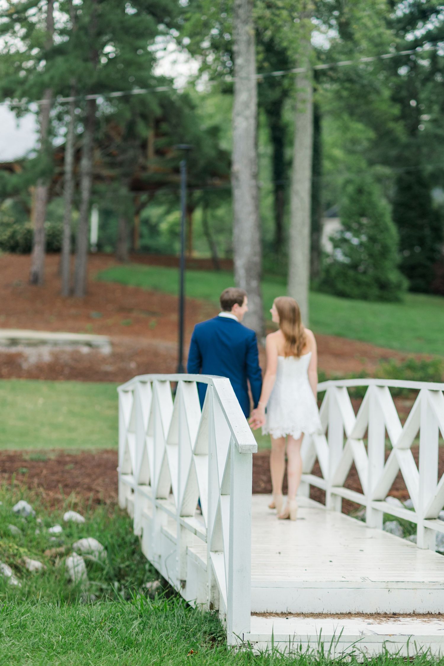 Couple holding hands walking across a white wooden bridge surrounded by lush green trees.
