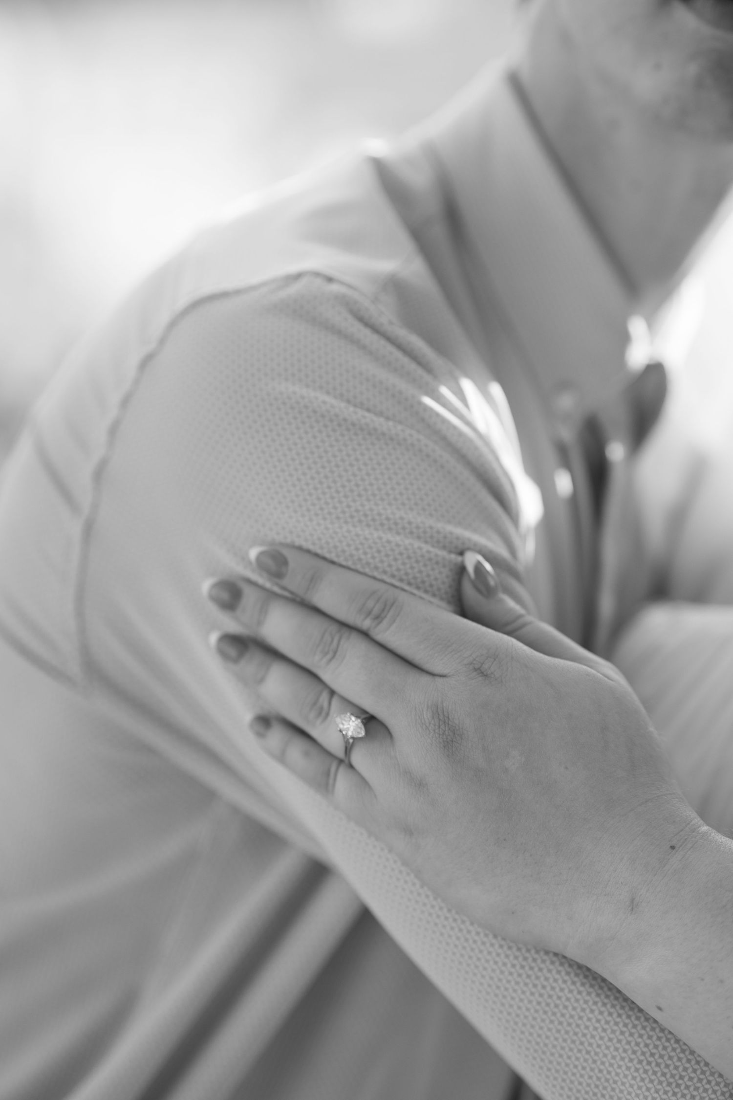 Close-up black and white photo of a woman's hand with a diamond engagement ring resting on a man's arm.