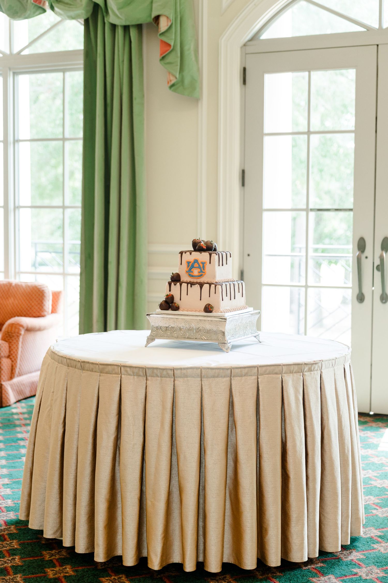 Elegant three-tier wedding cake with chocolate drizzle on a skirted table in a bright reception hall.