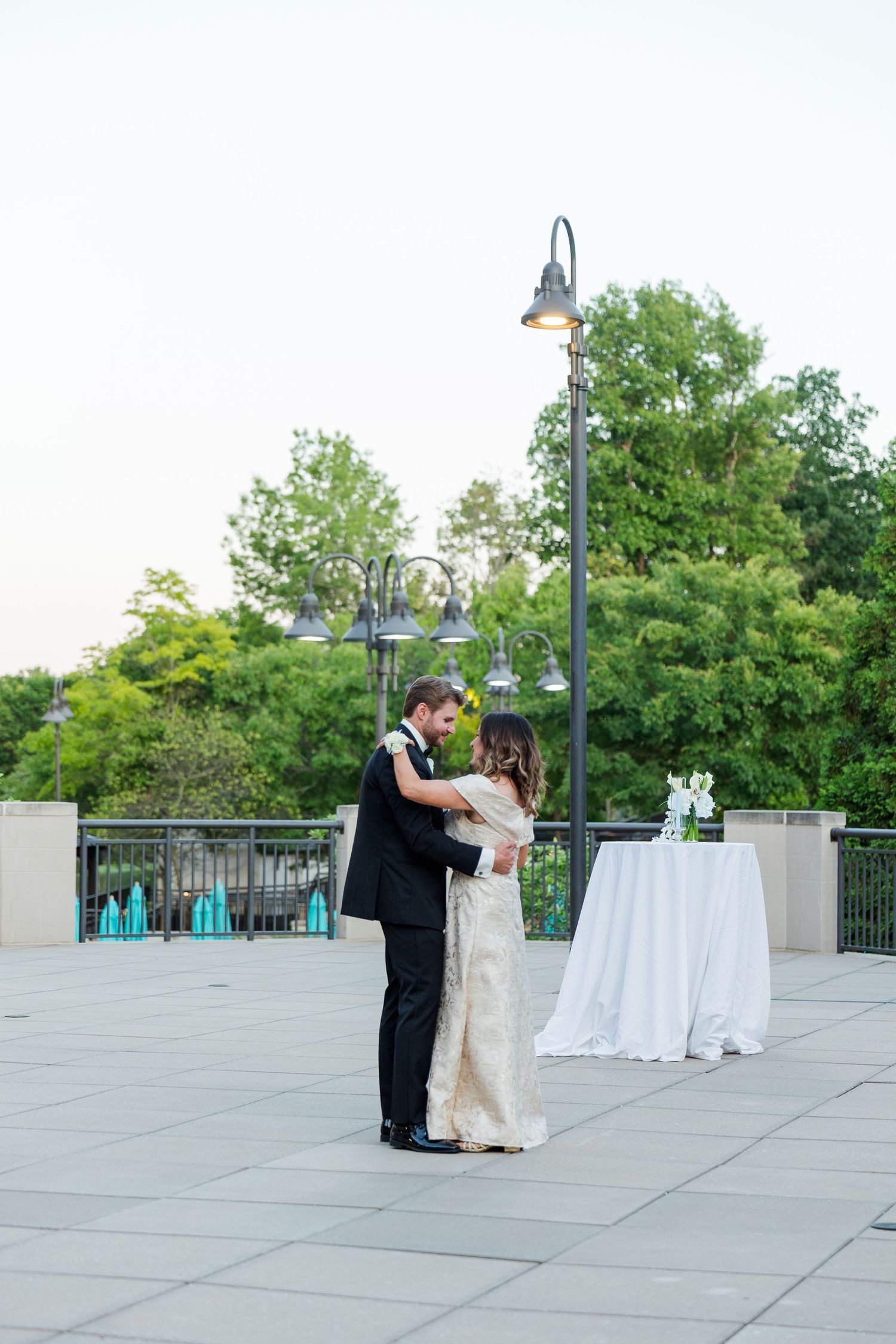 Bride and groom share their first dance on an outdoor terrace at dusk, surrounded by lush greenery and elegant lamp posts.