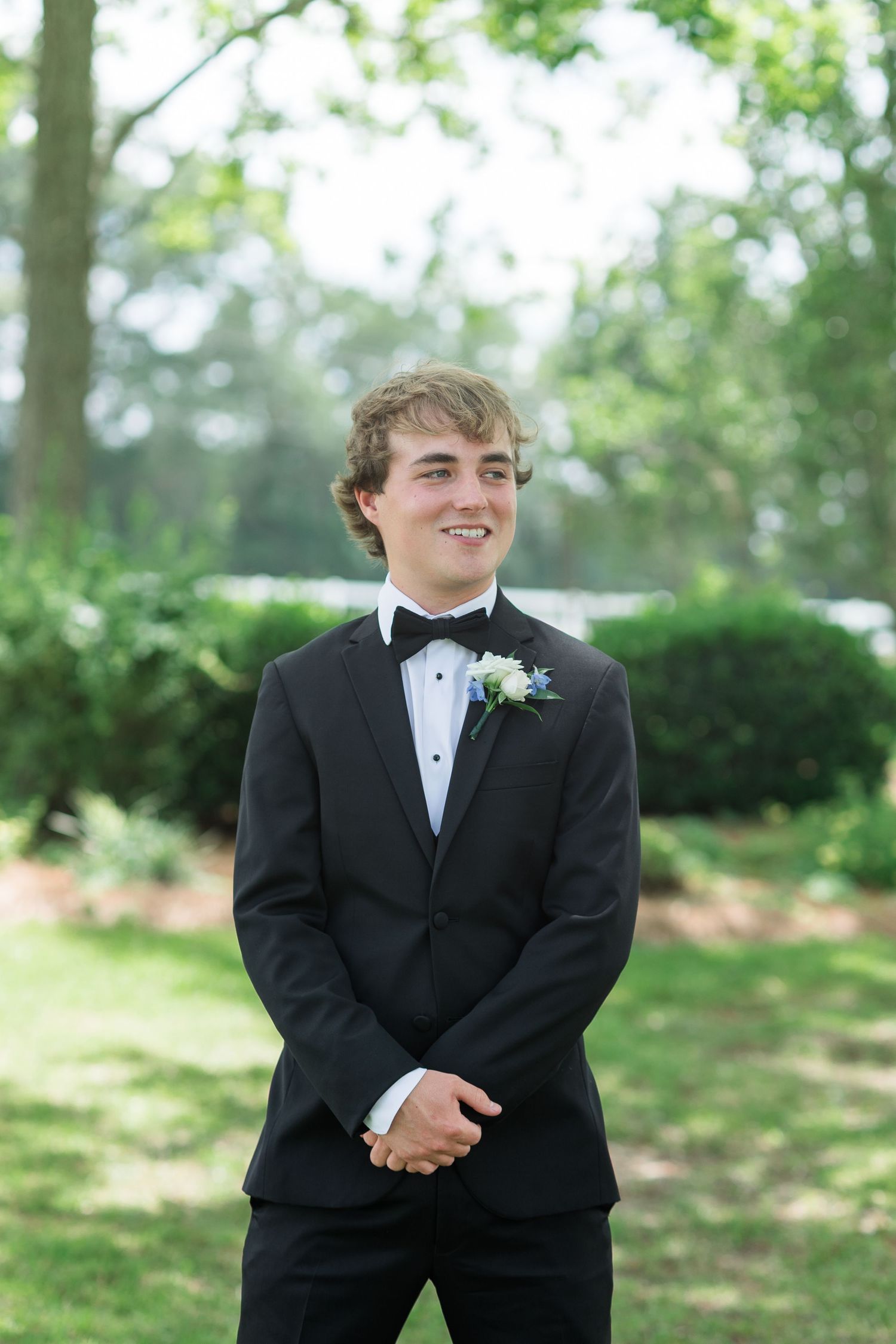 Young groomsman in black tuxedo with boutonniere smiles outdoors in lush green garden setting.