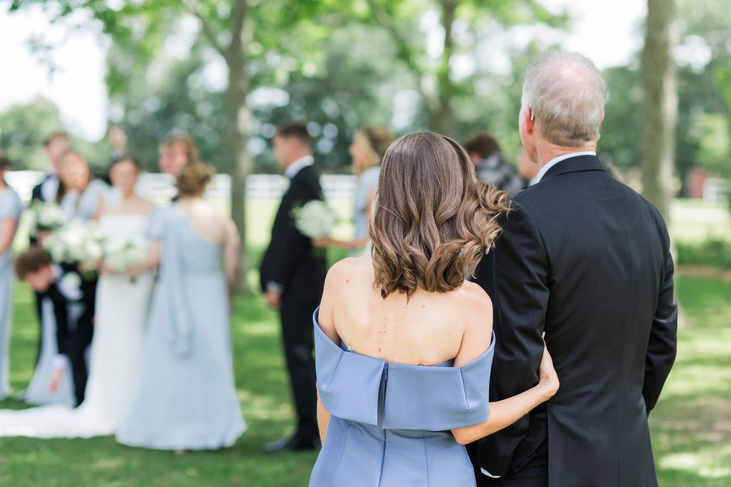 Mother and father of bride embrace from behind watching outdoor wedding ceremony with bridal party in background.