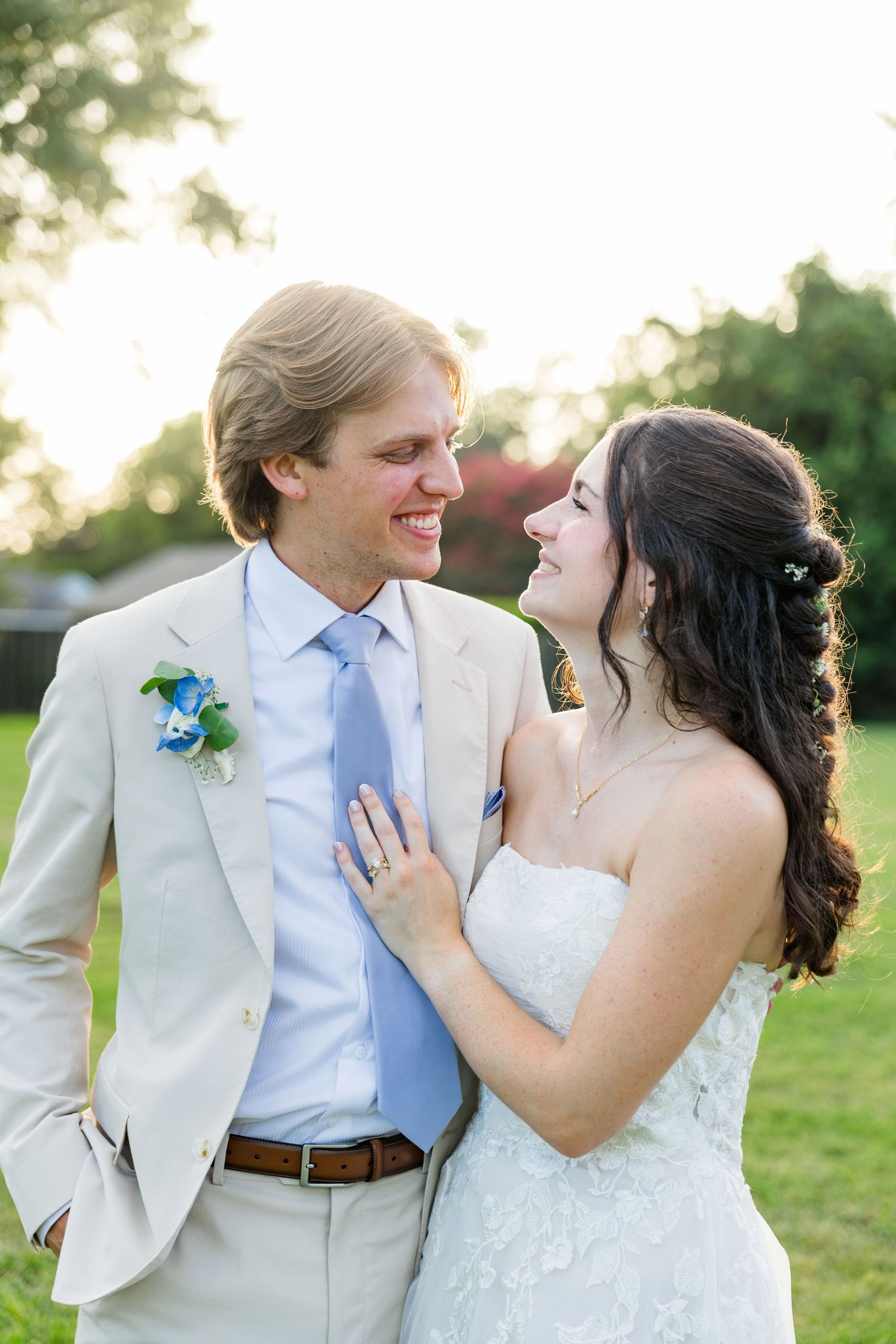 A smiling bride and groom share a romantic moment outdoors at golden hour, with the groom in a tan suit and blue tie.