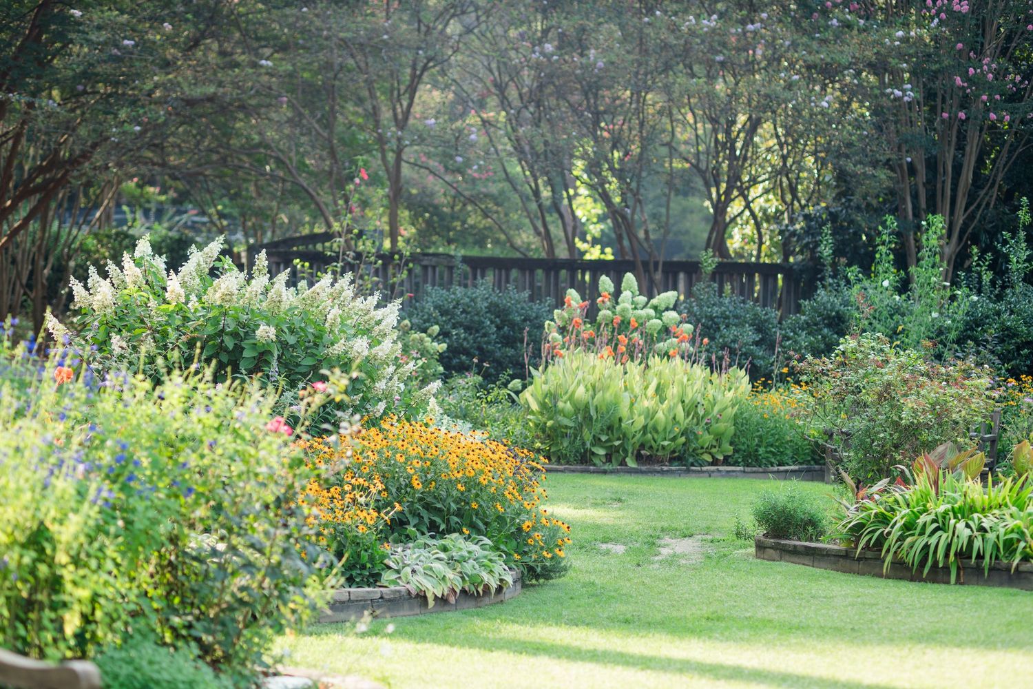 Lush garden with colorful flower borders, green lawn, and trees in background on a sunny day.