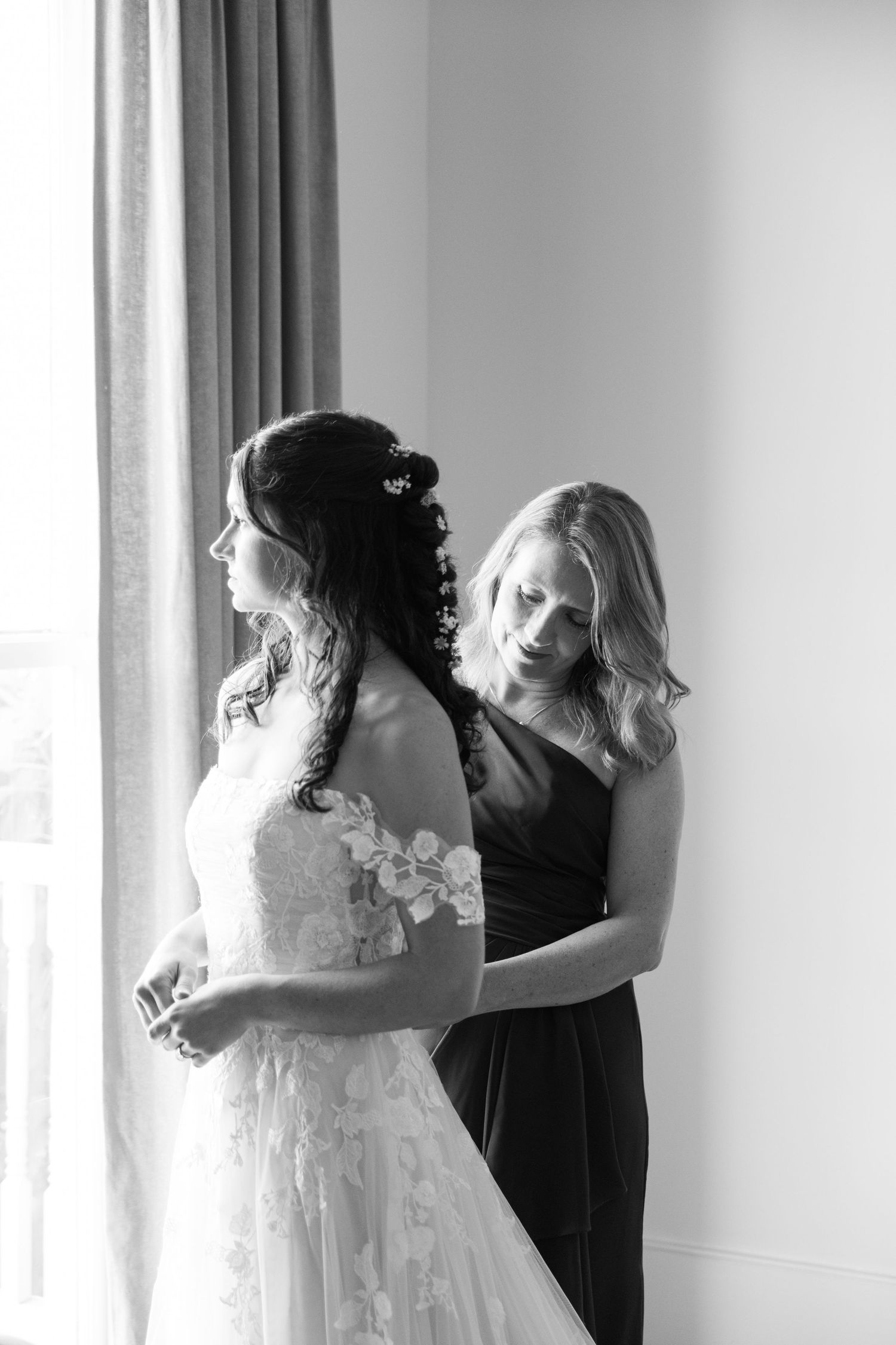 Black and white photo of bridesmaid zipping up bride's lace wedding dress by window.