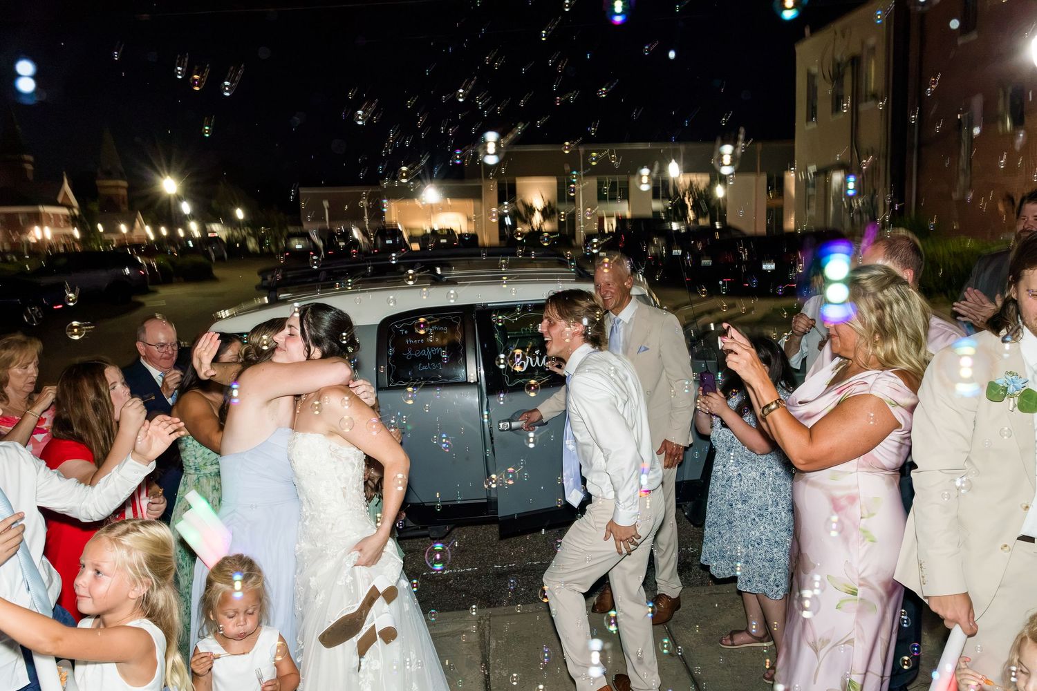 Newlyweds kissing outside at night surrounded by guests and bubbles near vintage getaway car.