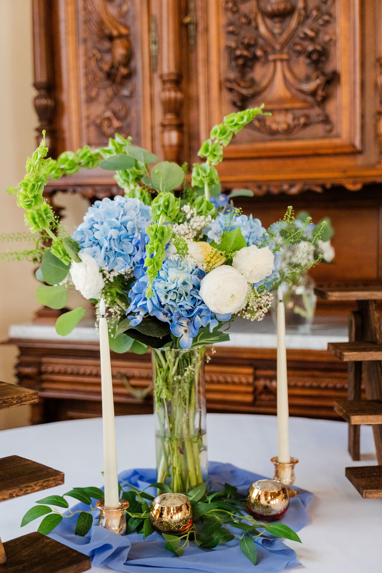 Blue hydrangea and white rose floral centerpiece with green bells of Ireland and taper candles on a white table.