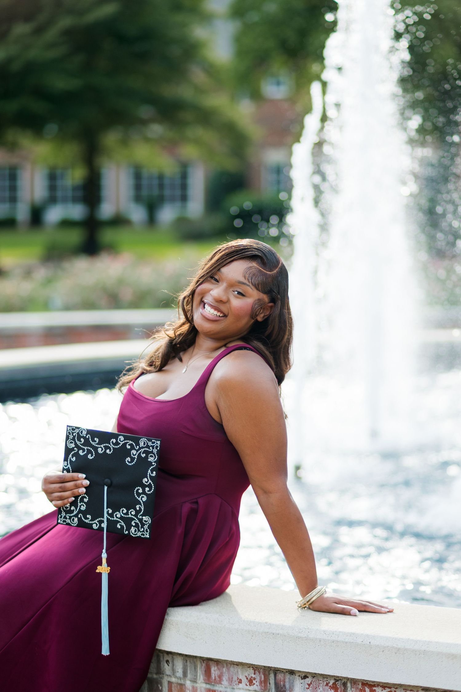 Smiling graduate in maroon gown holds decorated cap by fountain outdoors.