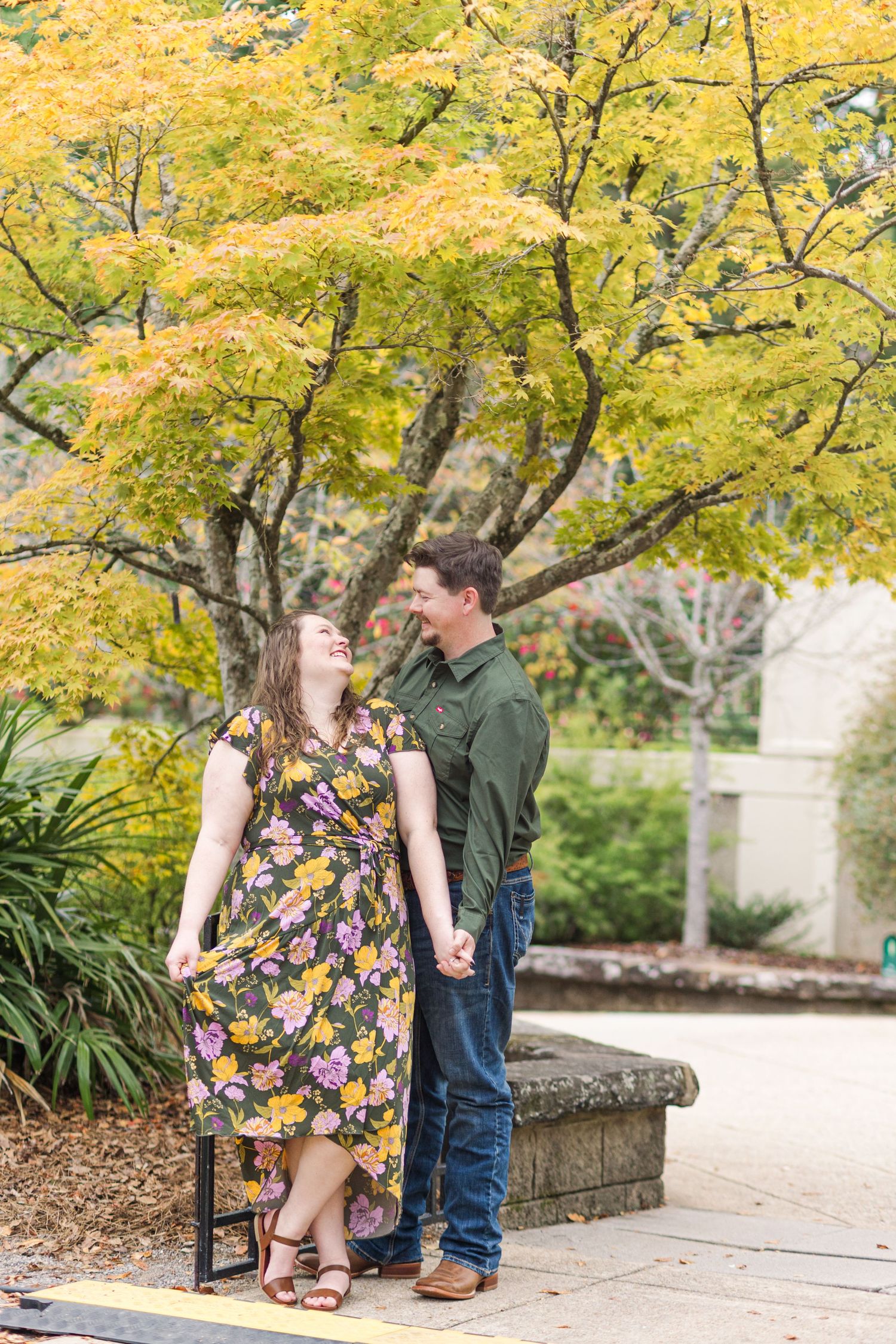 Couple holding hands laughing together outdoors in fall, woman in floral dress, man in green shirt, yellow autumn tree backdrop.