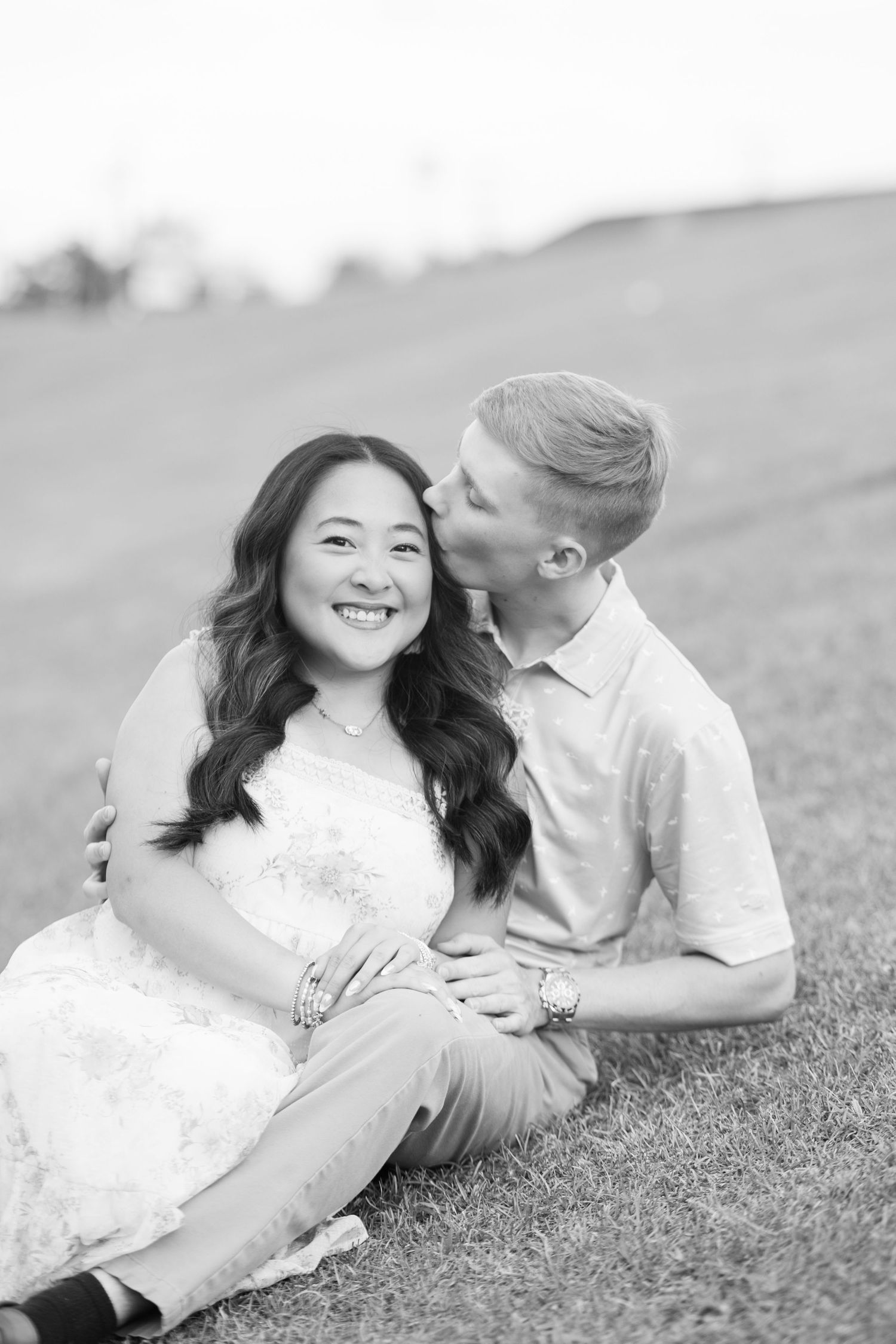 Black and white photo of a smiling woman and man sitting on grass, sharing a tender kiss on the cheek outdoors.