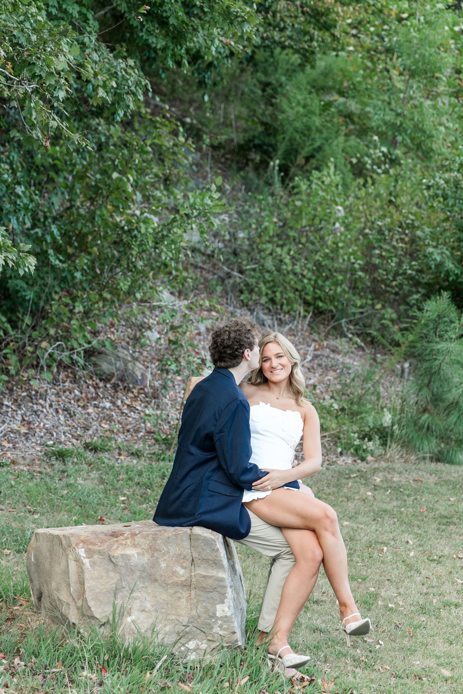 Couple sitting on a stone bench outdoors, man kissing woman's cheek as she smiles at the camera.