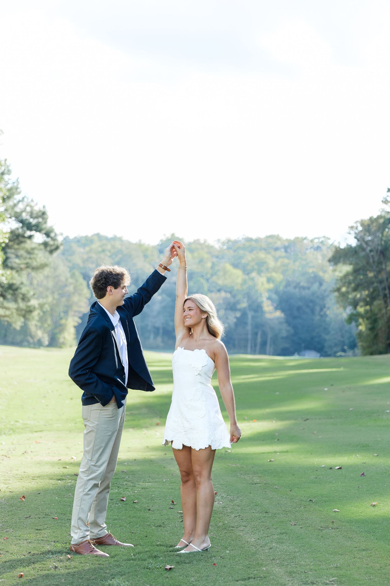 Happy couple dancing outdoors on a golf course green, man spinning woman in a white dress on a sunny day.