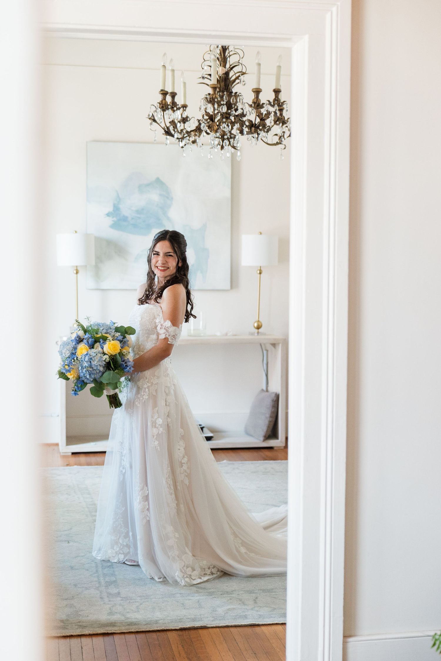 Smiling bride in a lace off-shoulder wedding gown holding a blue and yellow floral bouquet in an elegant white room.