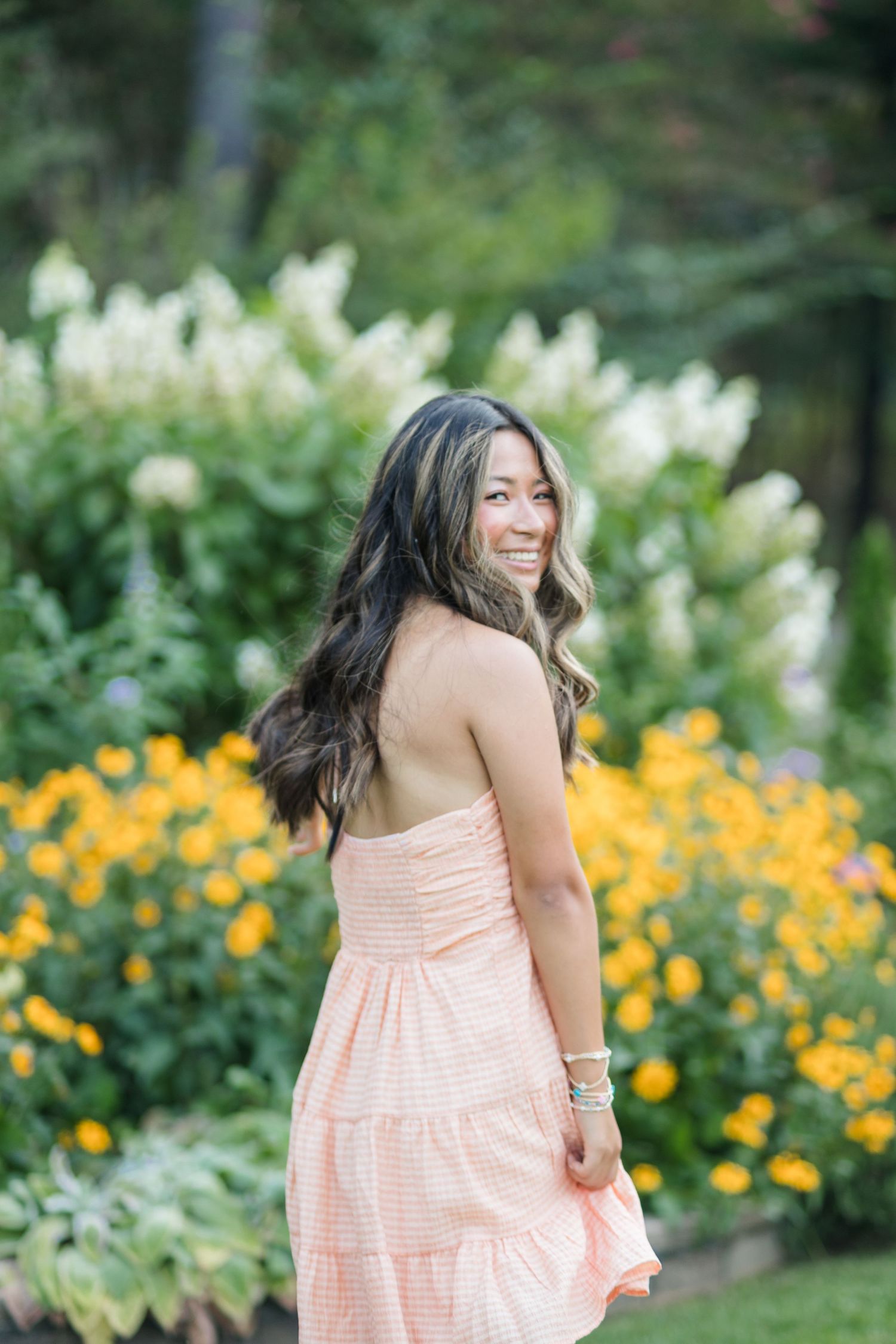 Young woman in strapless peach tiered dress smiling over shoulder in garden with yellow flowers.