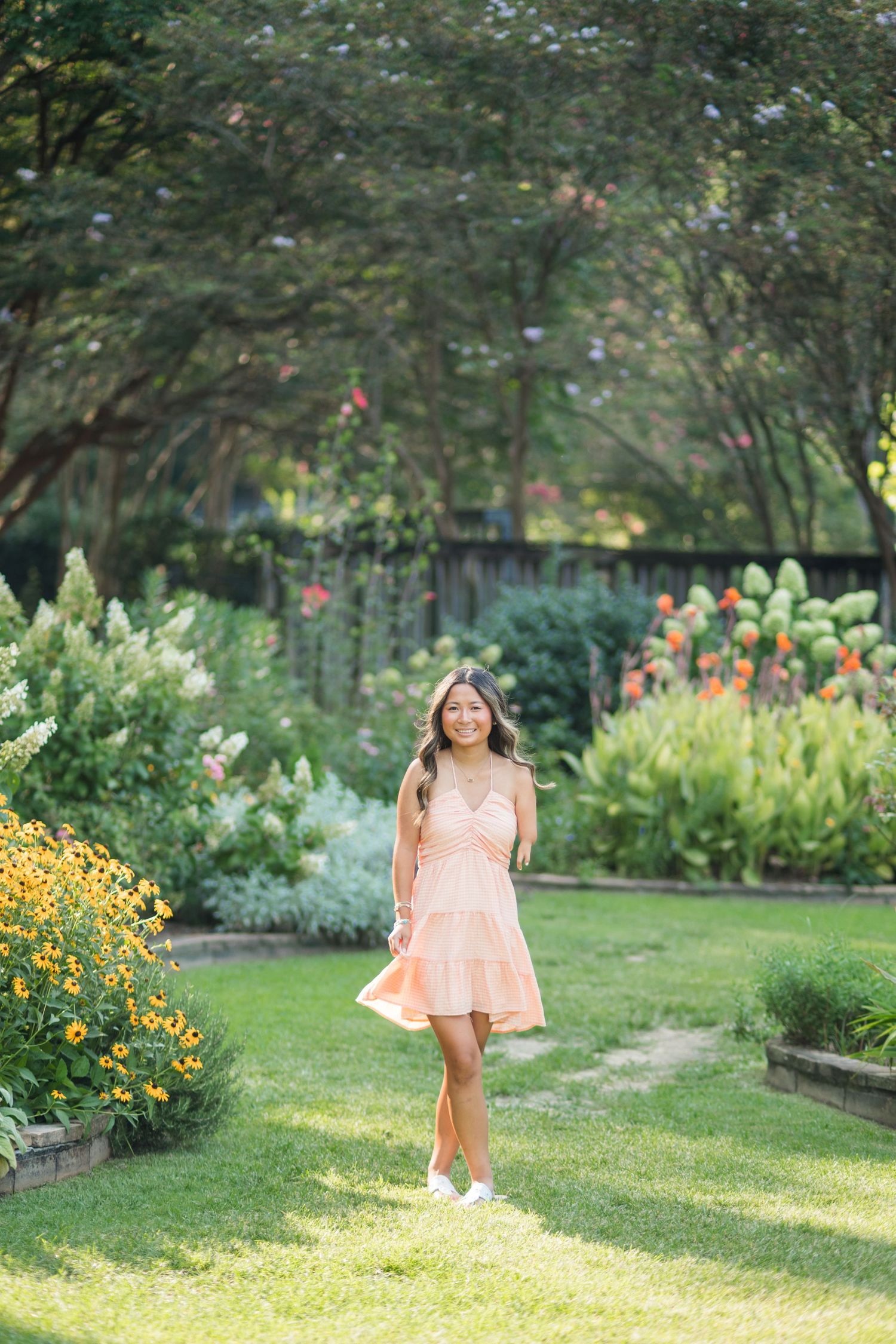 Young woman in peach dress walking through lush garden with colorful flowers and green trees on sunny day.