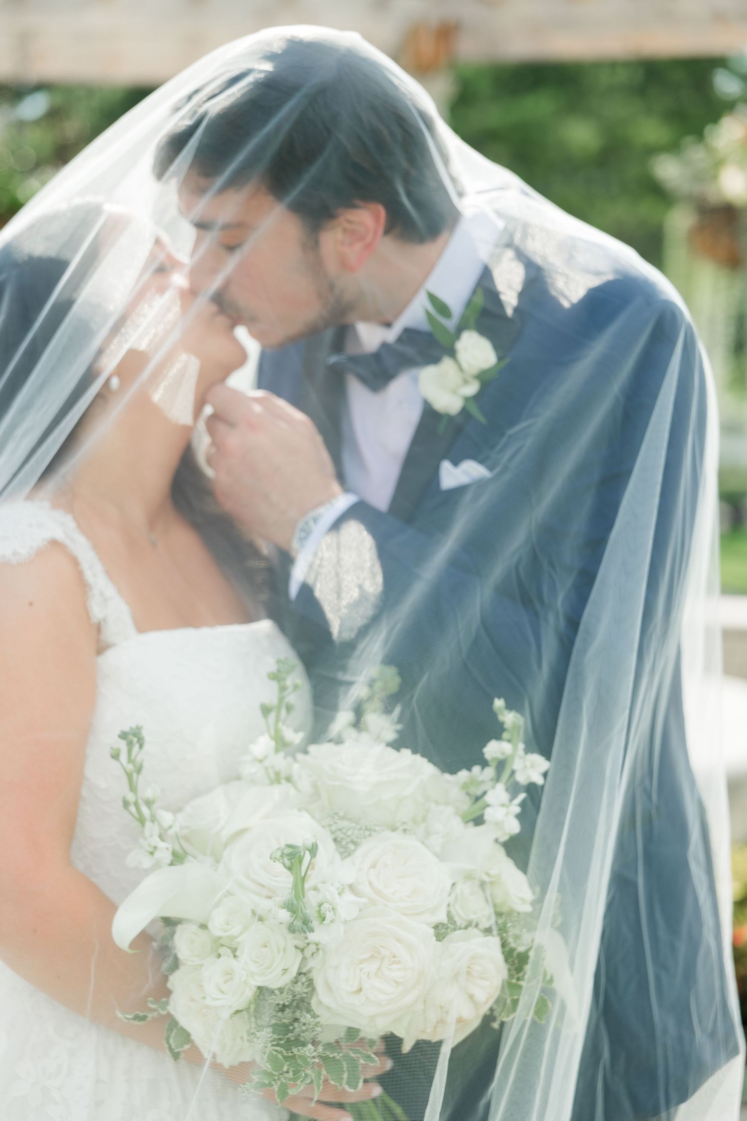 Bride and groom sharing a romantic kiss under a flowing wedding veil outdoors.