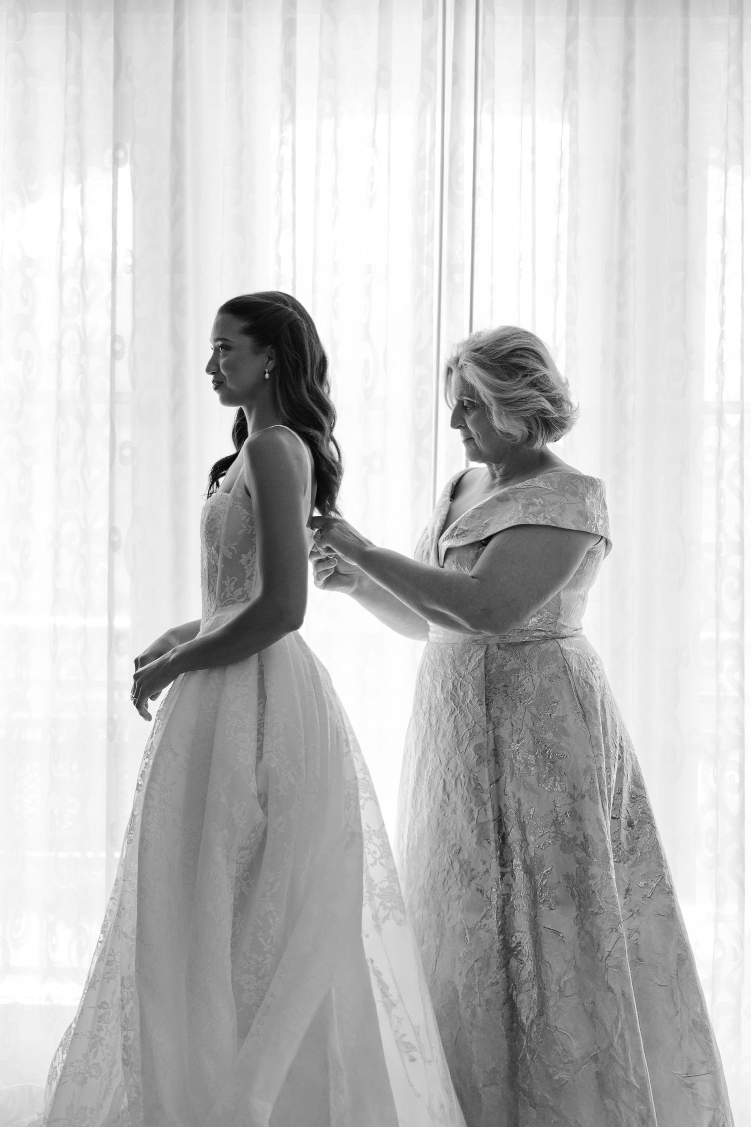 Black and white photo of a bride having her wedding dress fastened by an older woman in front of sheer curtains.