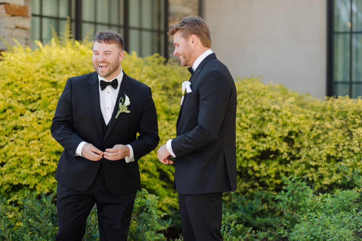 Two groomsmen in black tuxedos with white boutonnieres share a moment outdoors before a wedding ceremony.