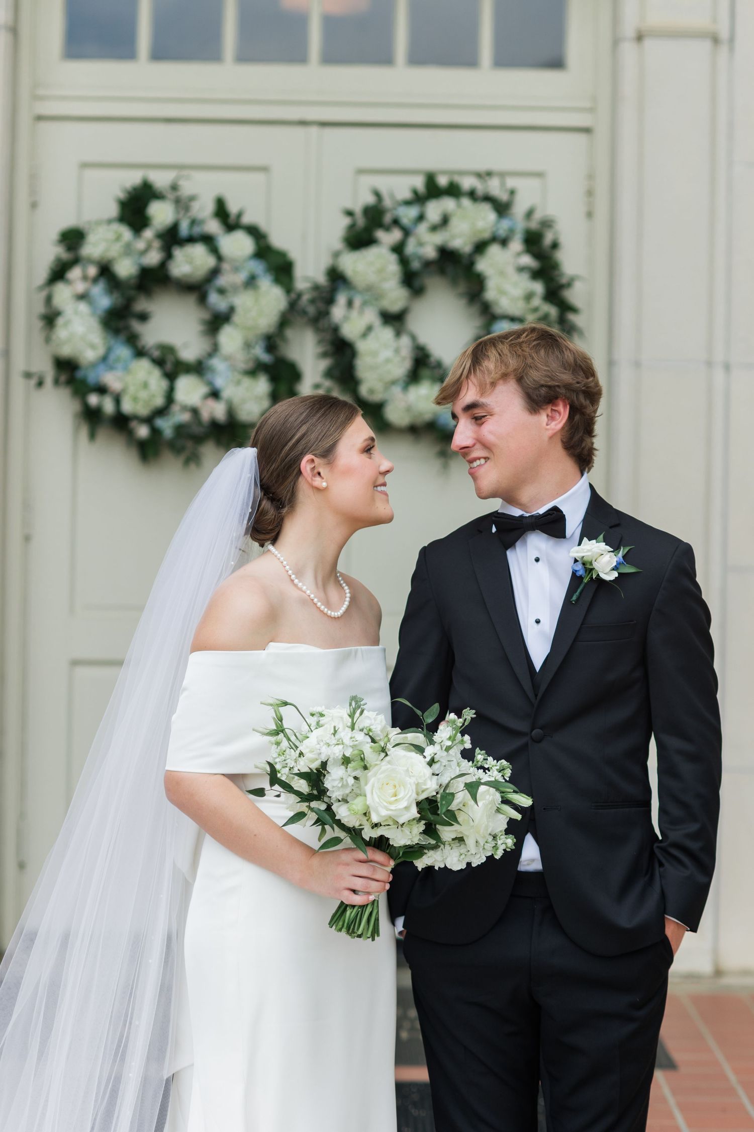 Bride and groom smiling at each other in front of a door adorned with white floral wreaths.