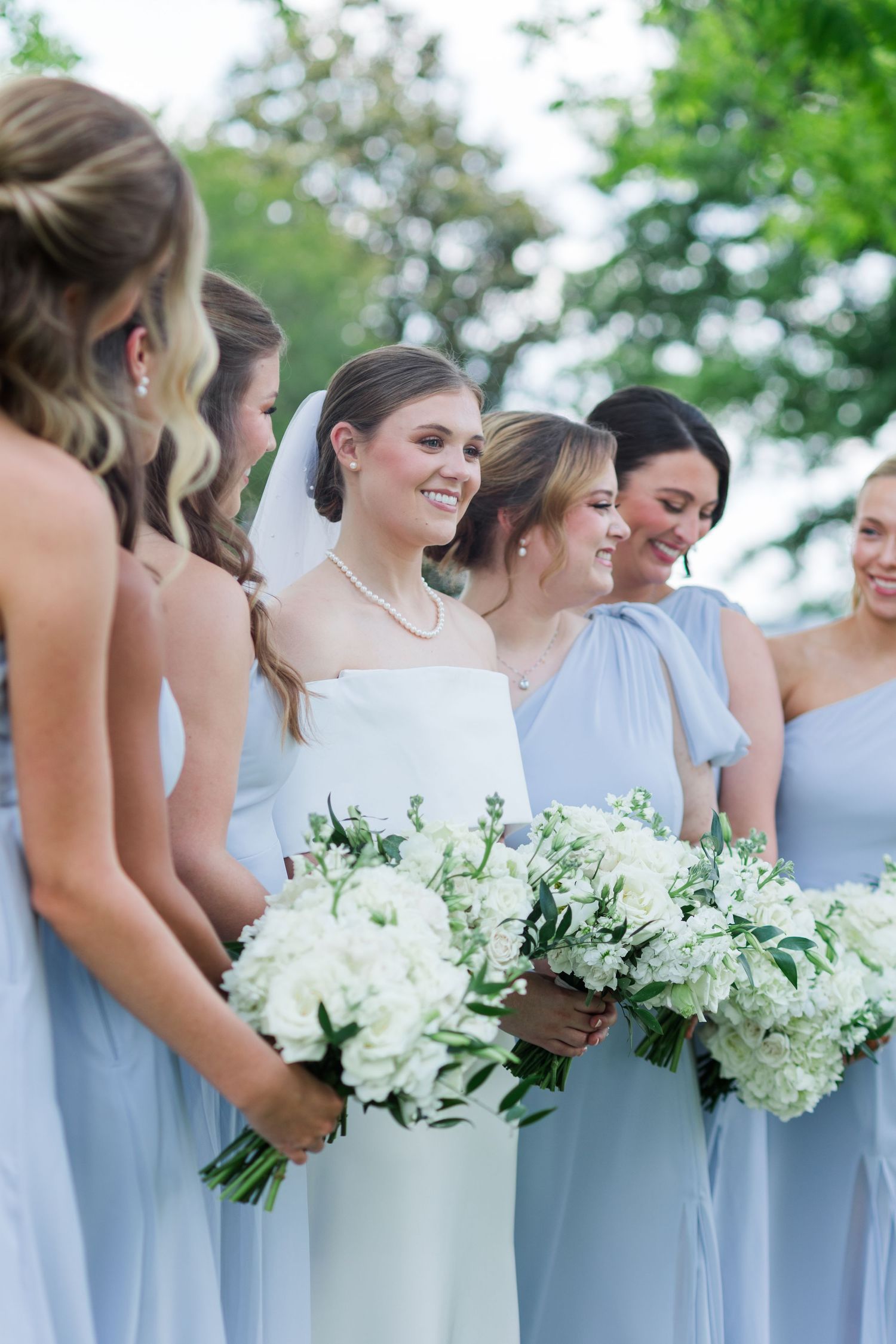 Smiling bride in white gown with pearl necklace stands among bridesmaids in light blue dresses holding white bouquets.