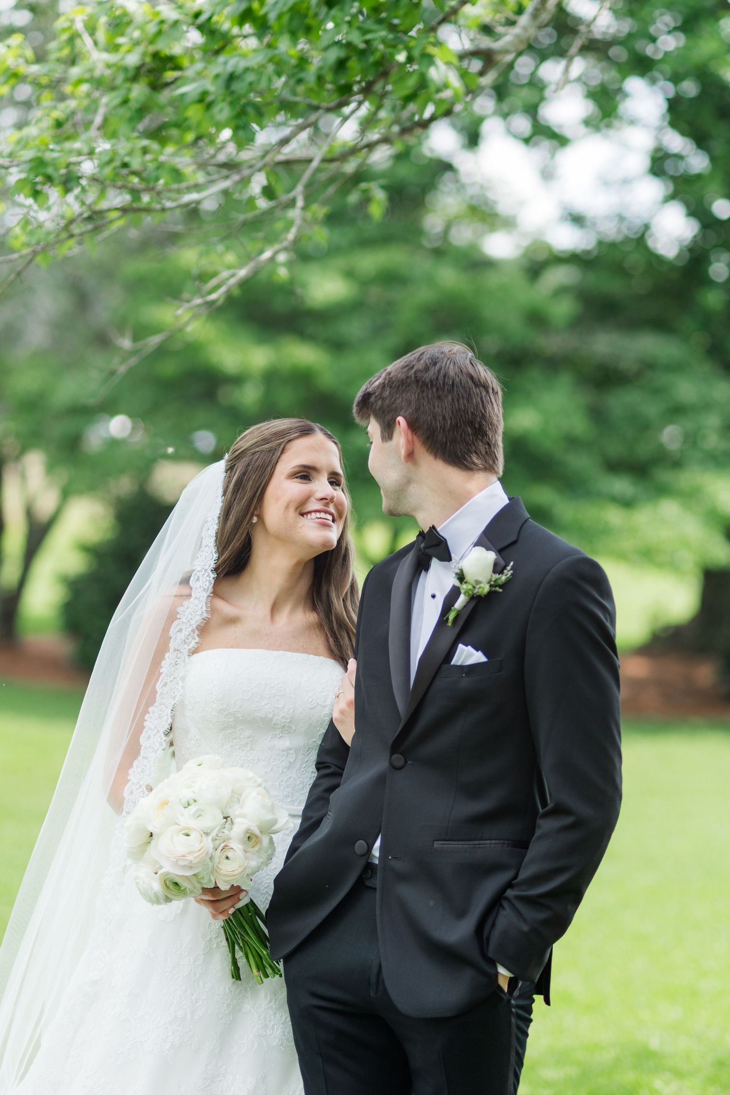 Smiling bride in white gown and veil holds bouquet while gazing at groom in black tuxedo outdoors.