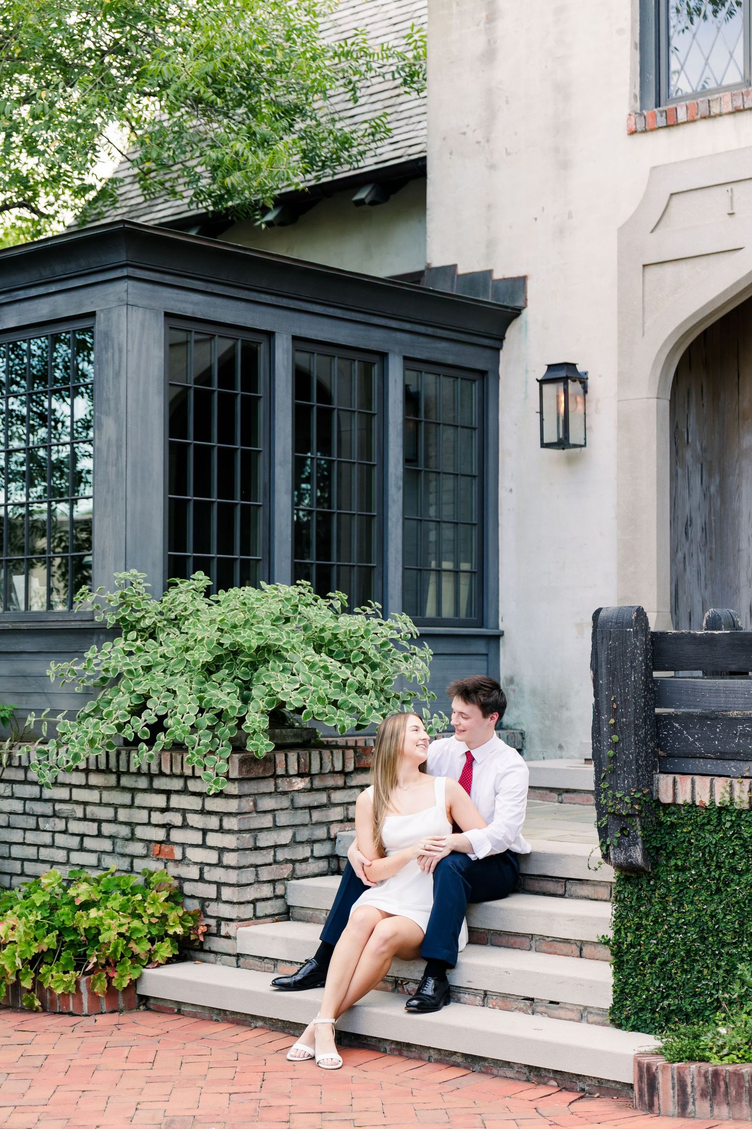 A couple sits together on brick steps outside an elegant home surrounded by lush greenery.