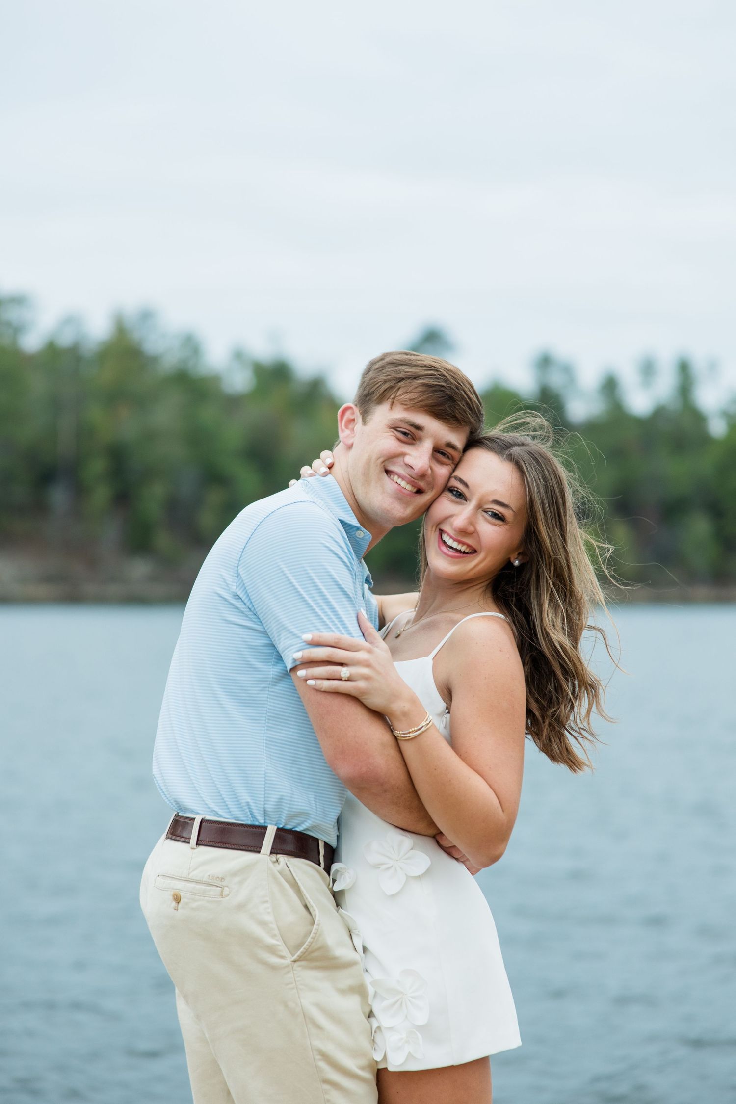Happy couple embracing and laughing in front of a lake with trees in the background, woman showing her ring.