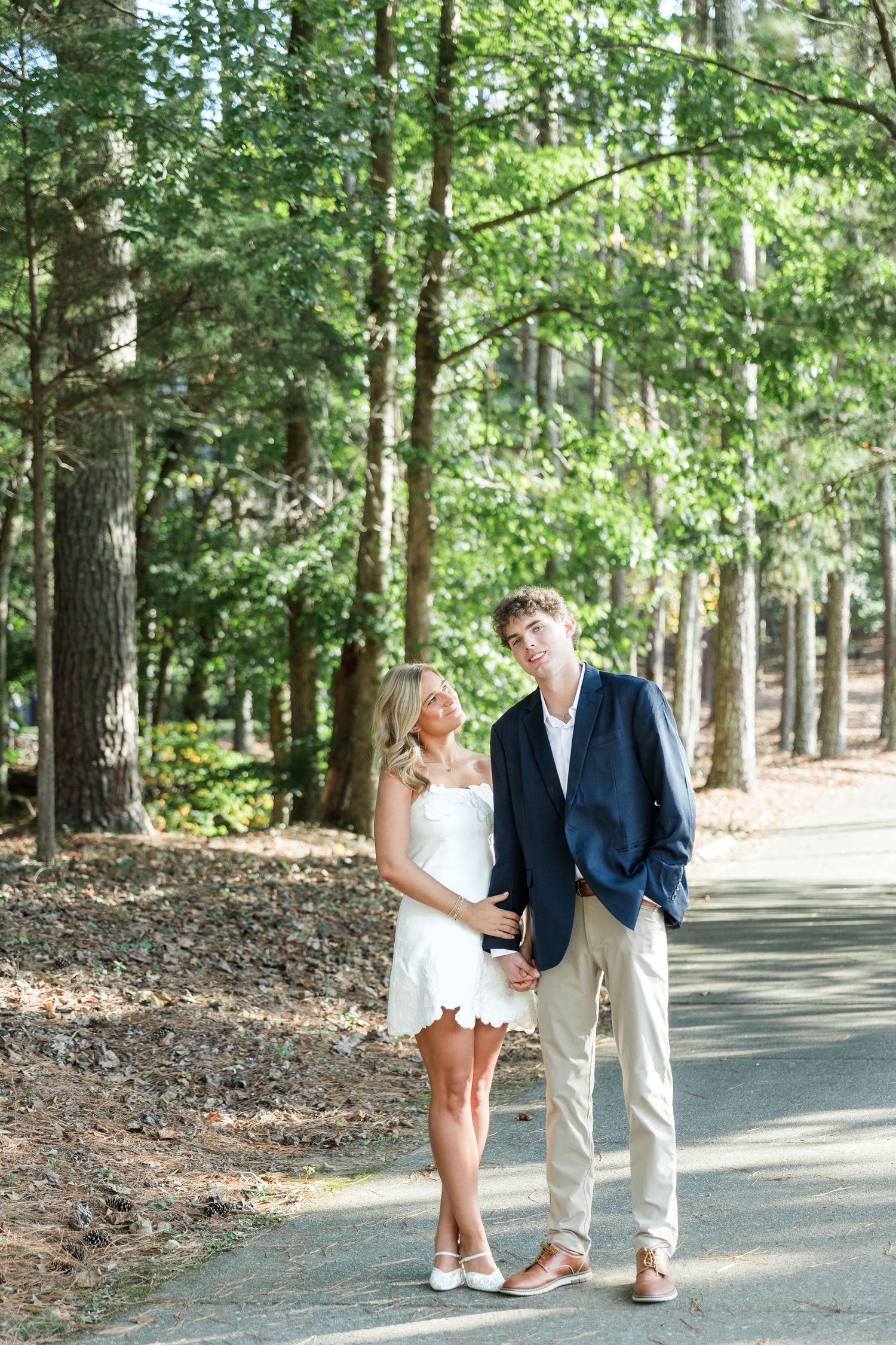 Couple standing together on a tree-lined path, woman in white dress looking up at tall man in navy blazer.
