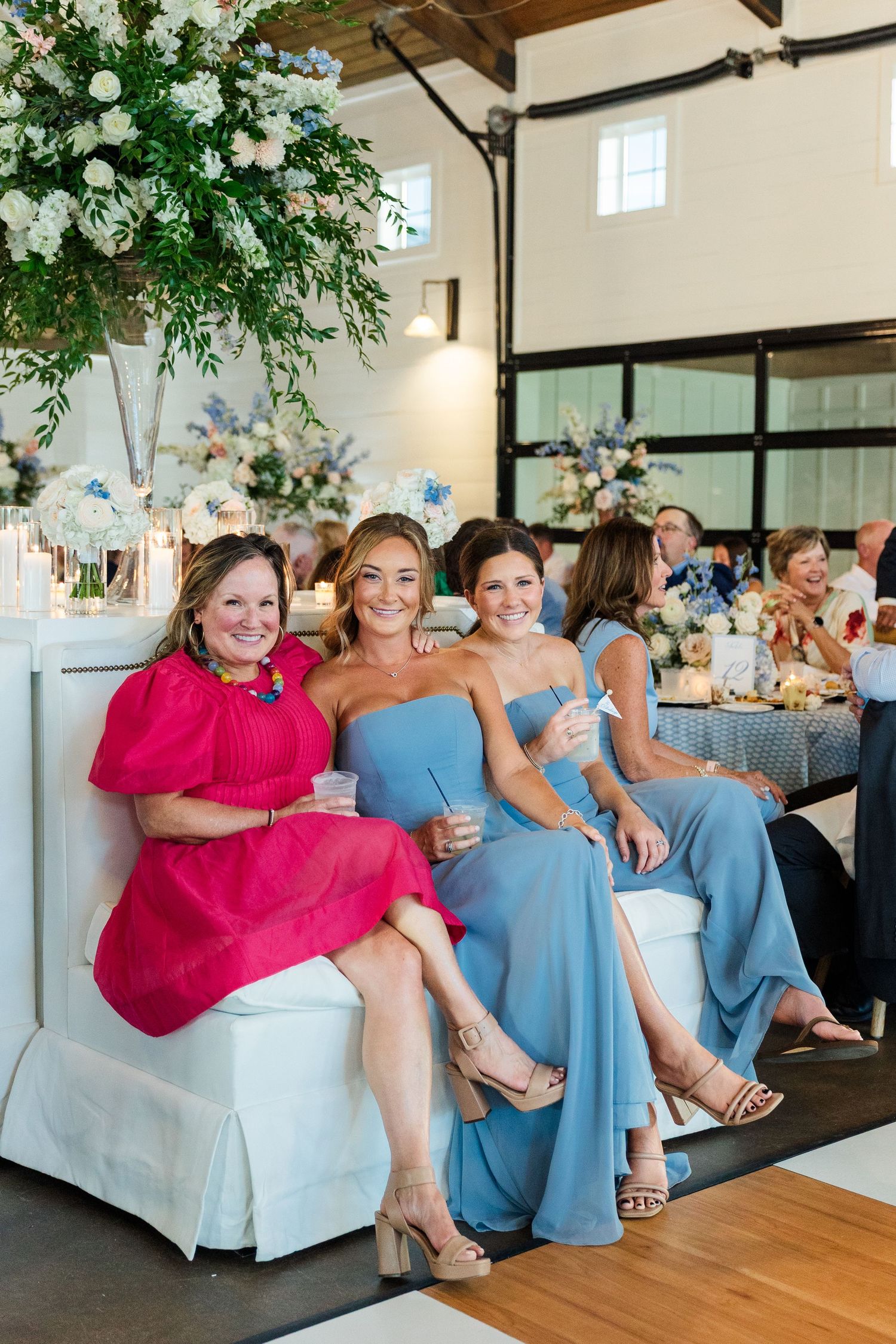 Three smiling women in elegant dresses pose together at a wedding reception with floral arrangements.