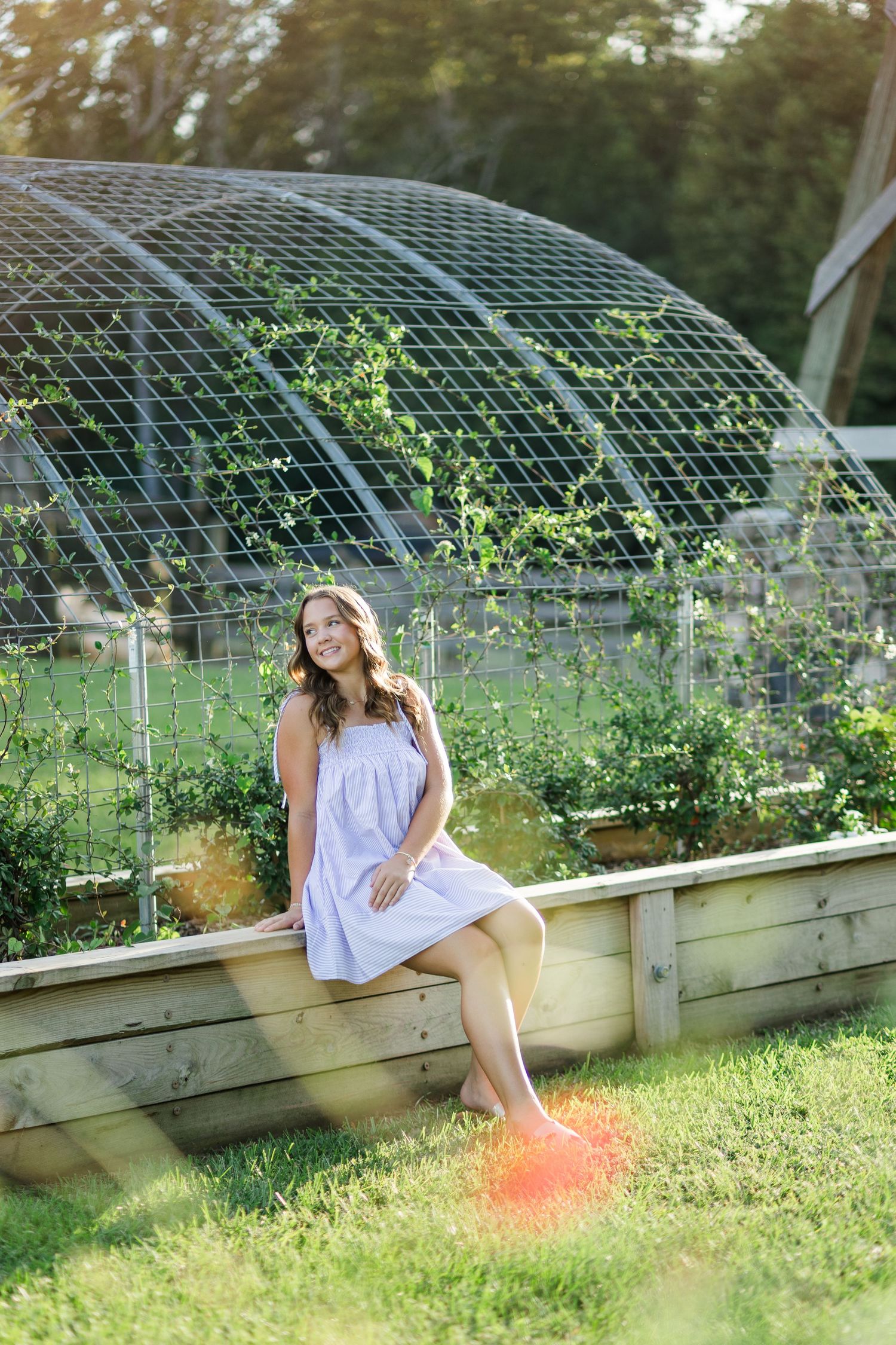 Young woman in white dress sitting on raised garden bed beside dome trellis in sunny outdoor garden setting.