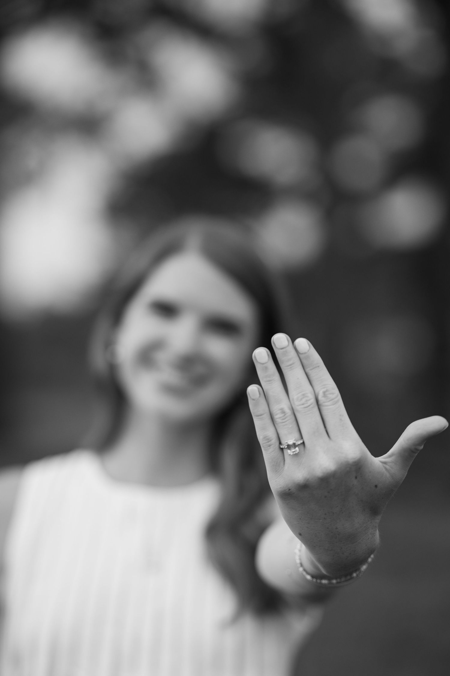 Black and white photo of smiling woman showing off engagement ring on outstretched hand toward camera.