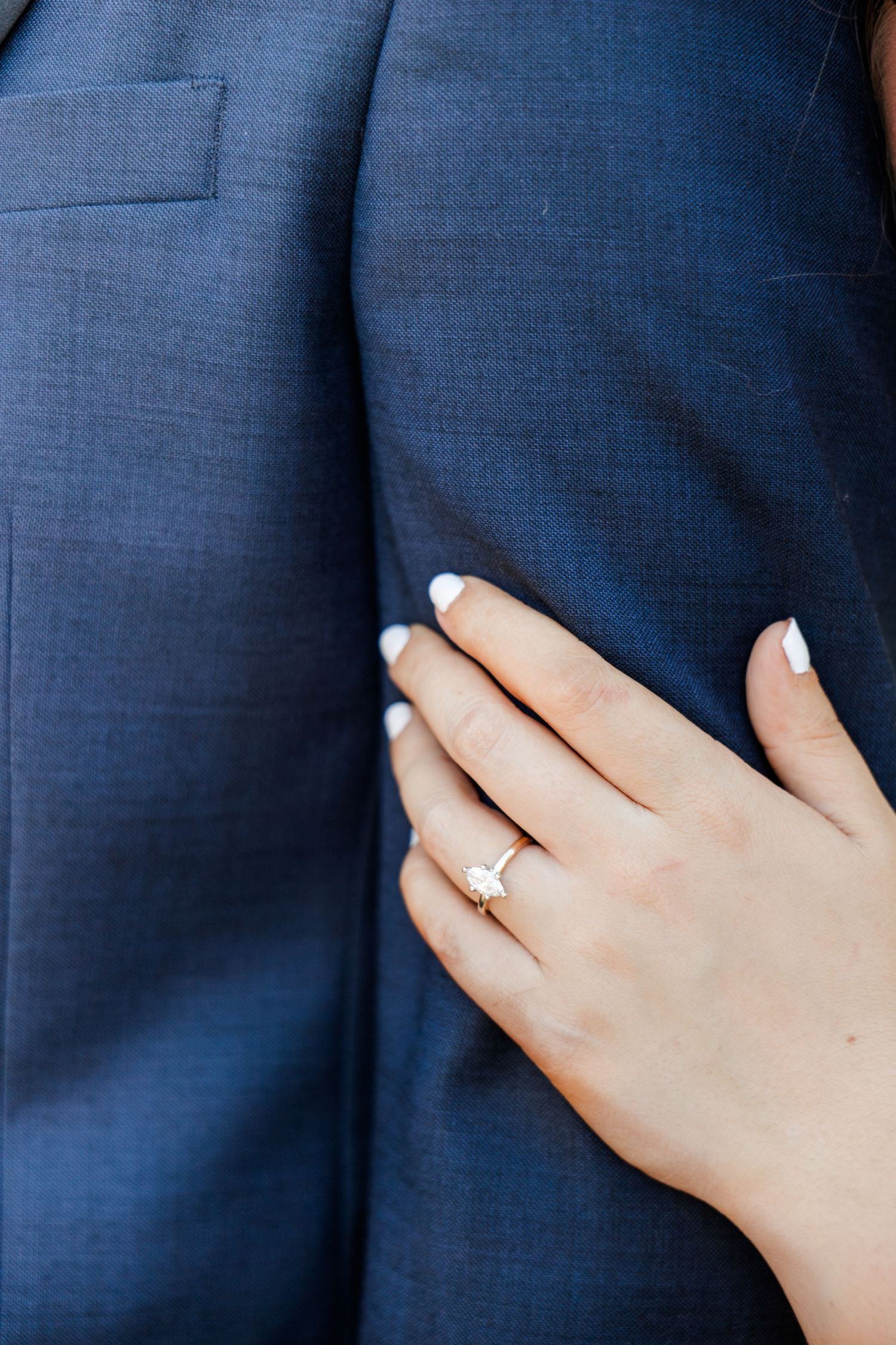 Close-up of a woman's hand with an engagement ring resting on a man's navy blue suit jacket.