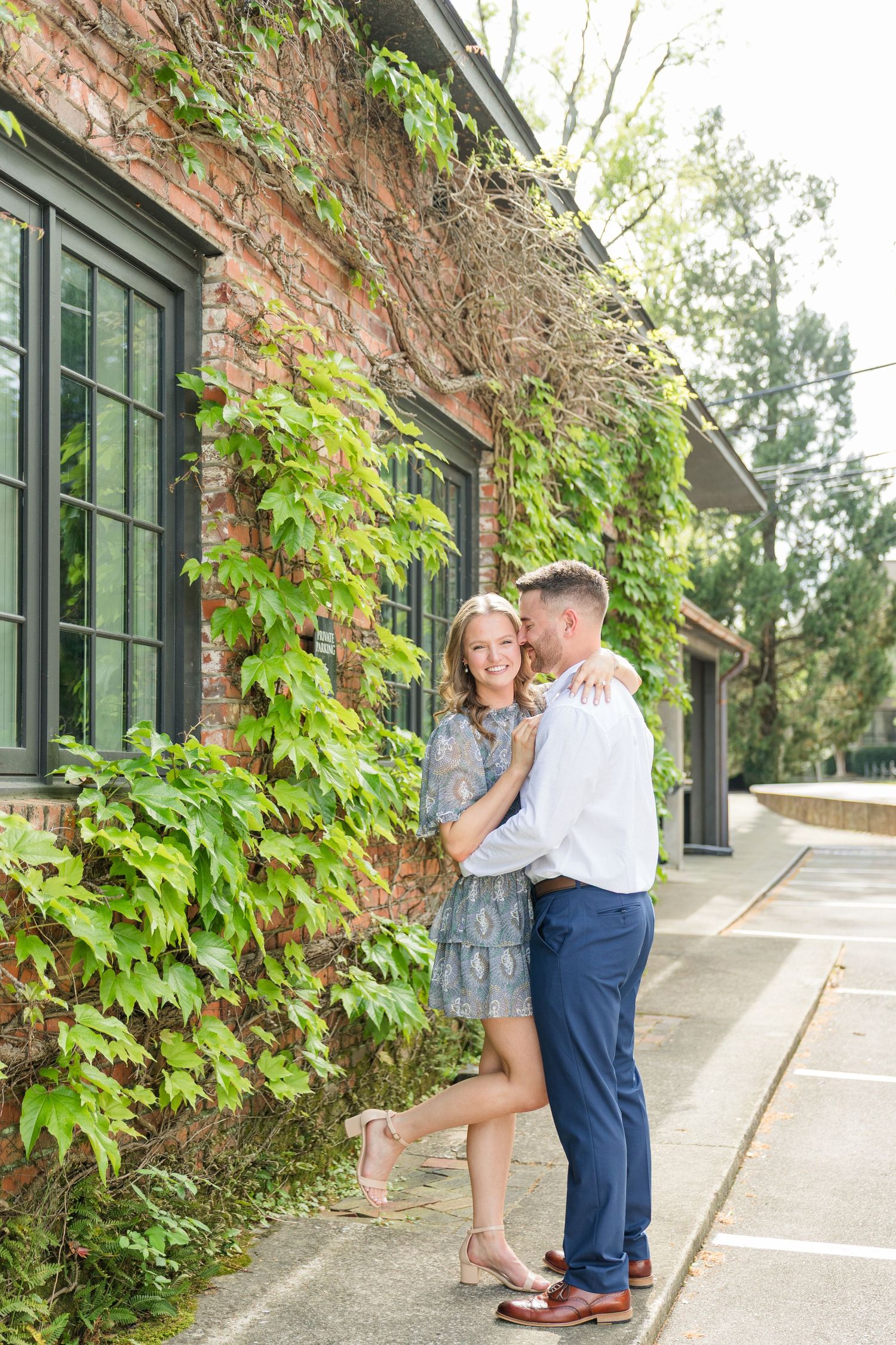 Couple embracing and smiling against ivy-covered brick wall with black windows outdoors.