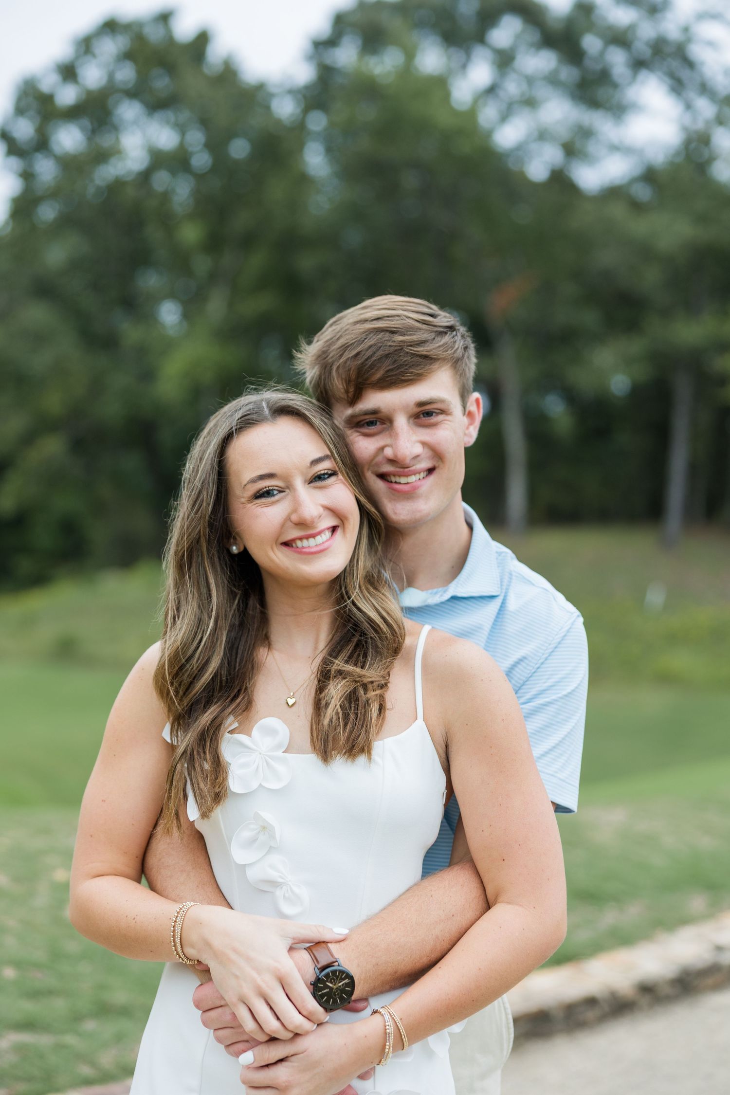 Happy engaged couple embracing outdoors, the woman in a white dress laughing while the man hugs her from behind near a tree-lined green.