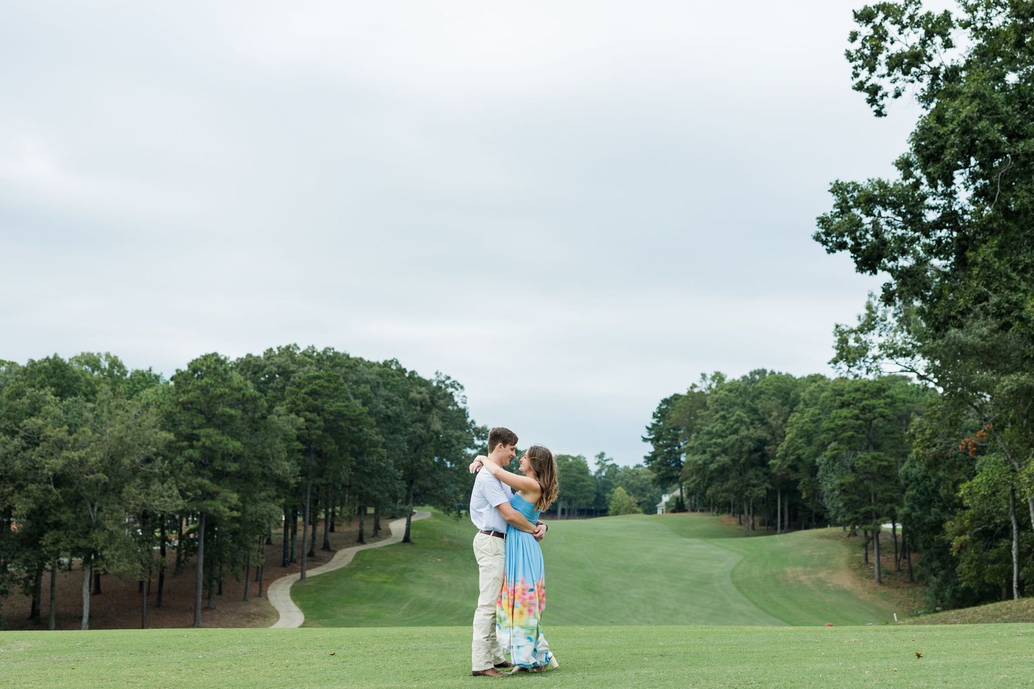 Engaged couple hugging on a green golf course with a curving path and tall trees framing the scene beneath a cloudy sky.