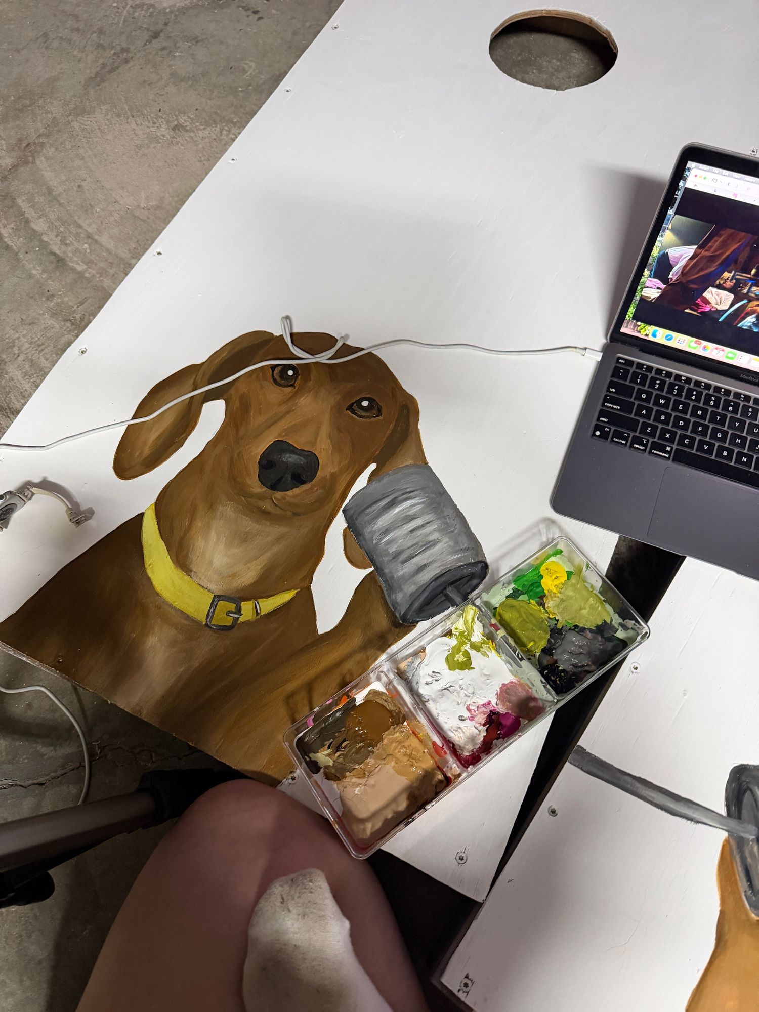 Artist painting a dachshund dog portrait on a canvas board with a laptop reference photo nearby.