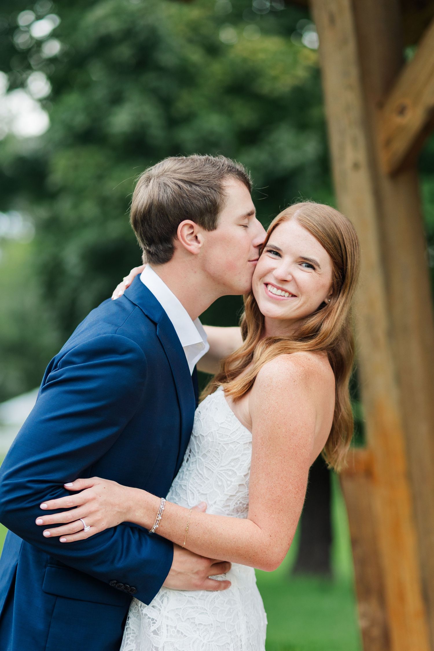 Groom in blue suit kisses bride on cheek as she smiles joyfully outdoors near wooden post.