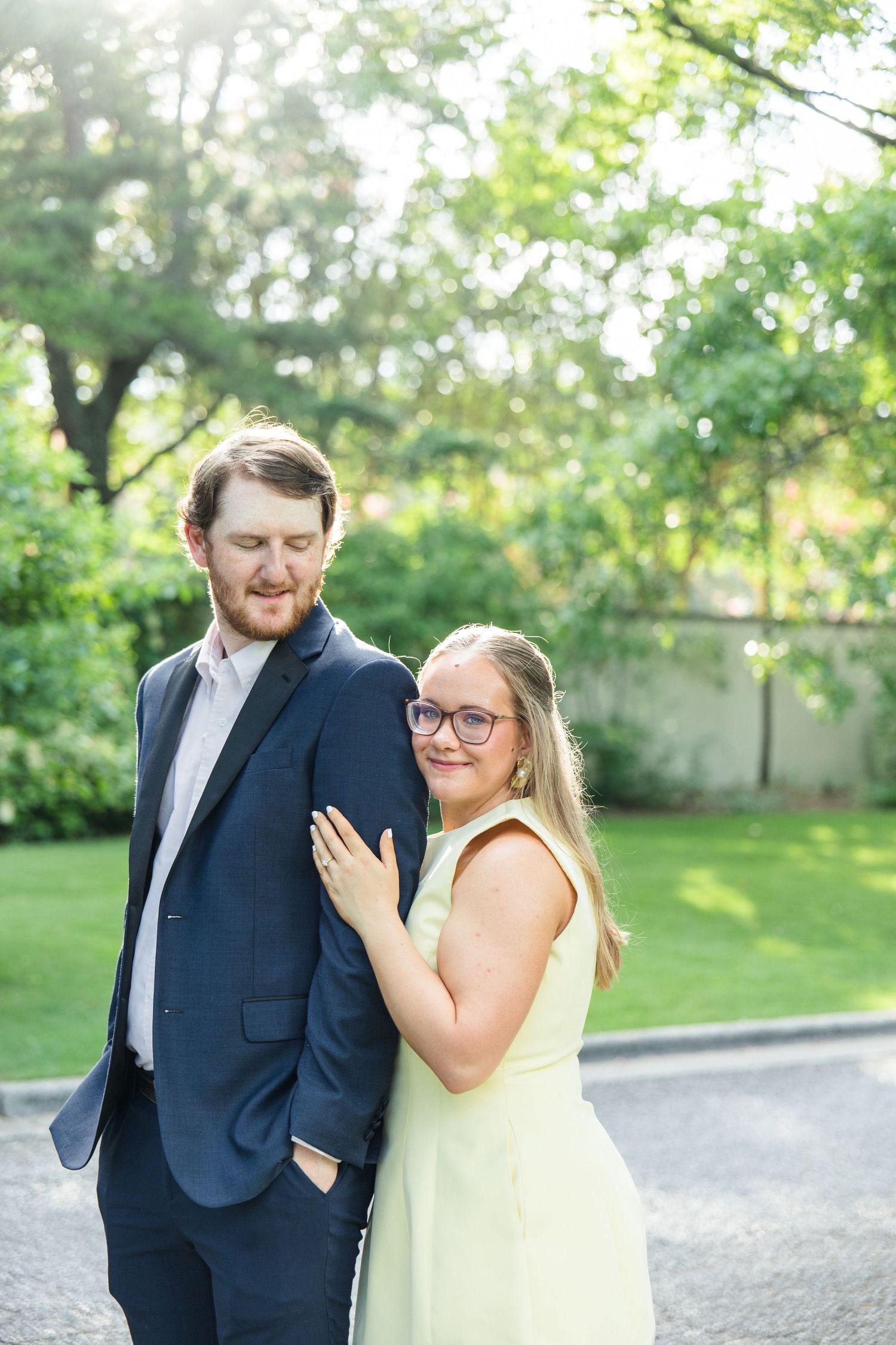 Couple posing outdoors, woman in light green dress leaning on man in navy suit, lush green trees in background.