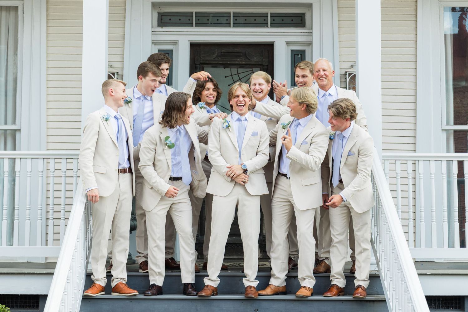 Groomsmen in tan suits and blue ties pose on white porch steps with groom at center for wedding photo.
