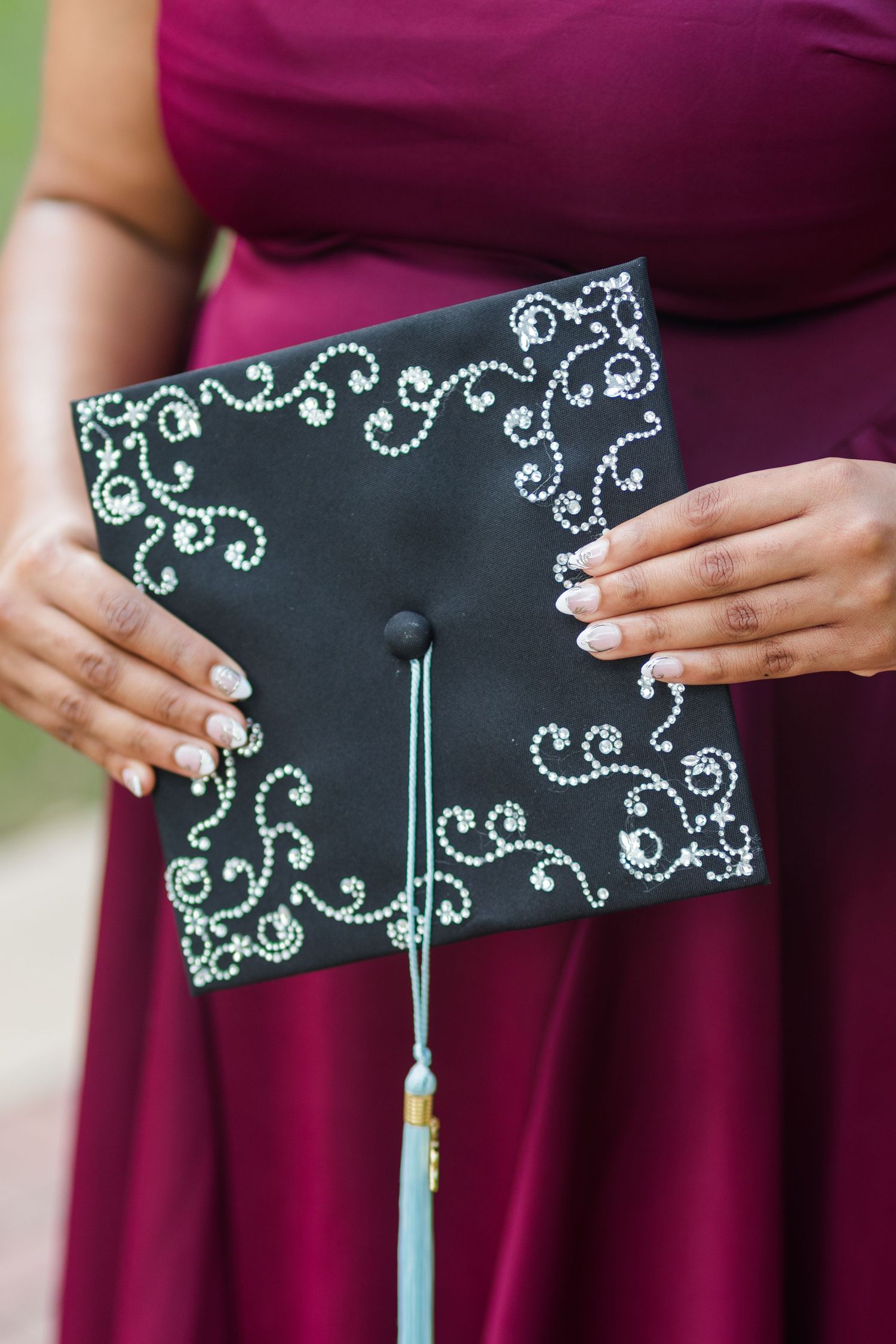 Person in burgundy dress holding decorated black graduation cap with silver swirl rhinestone designs.