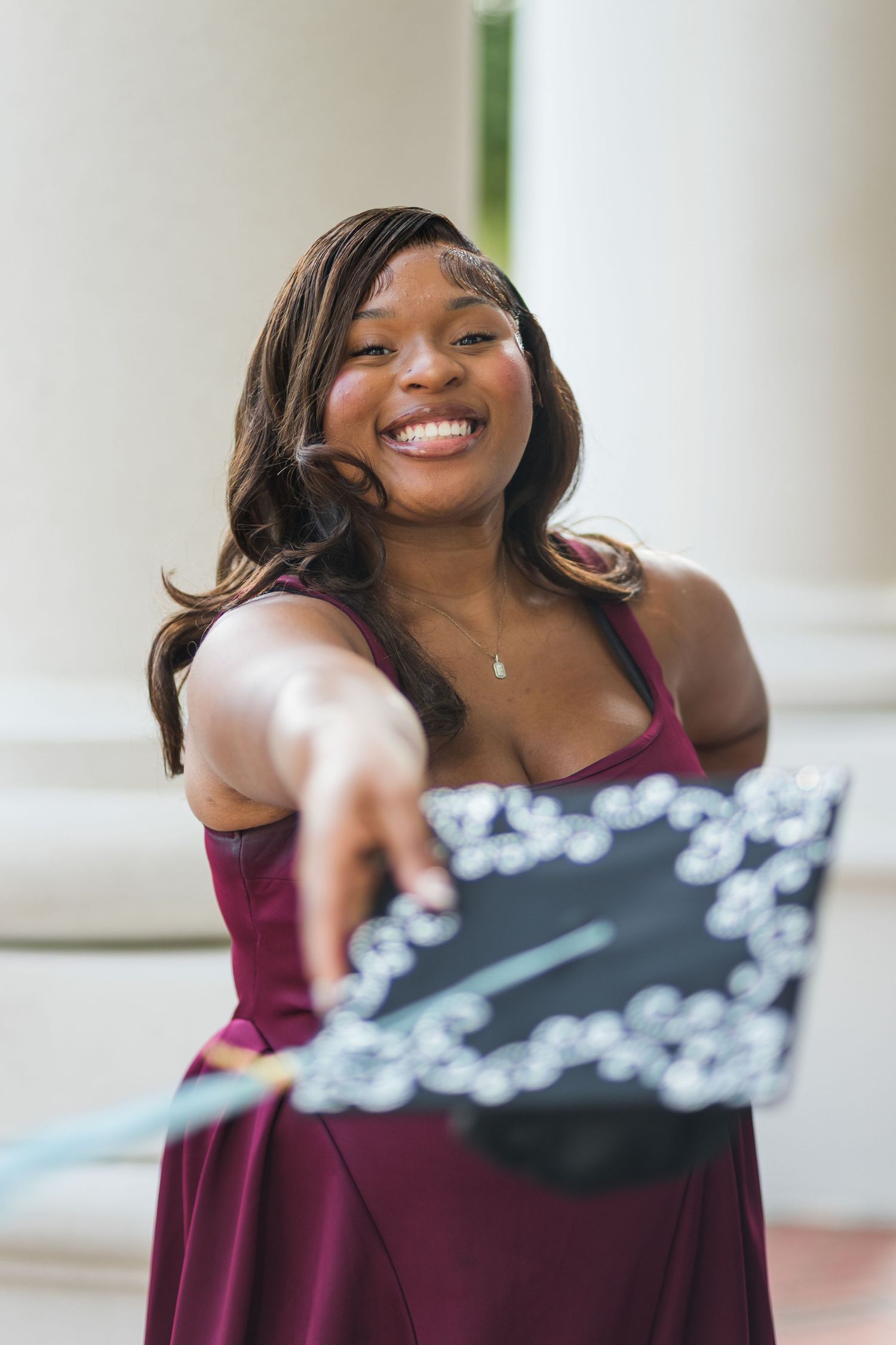 Smiling young woman in burgundy dress holding decorated graduation cap toward camera near white columns.
