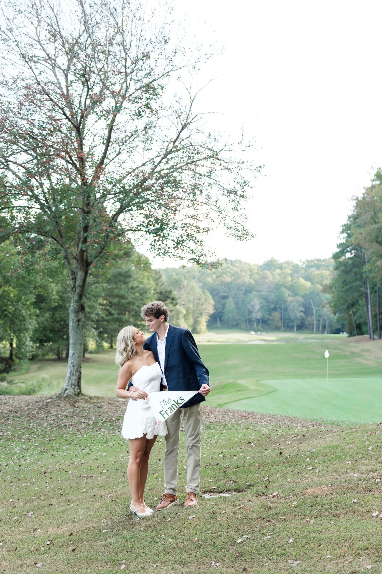 Romantic couple kissing on golf course, woman in white dress and man in navy blazer holding a sign in lush green setting.