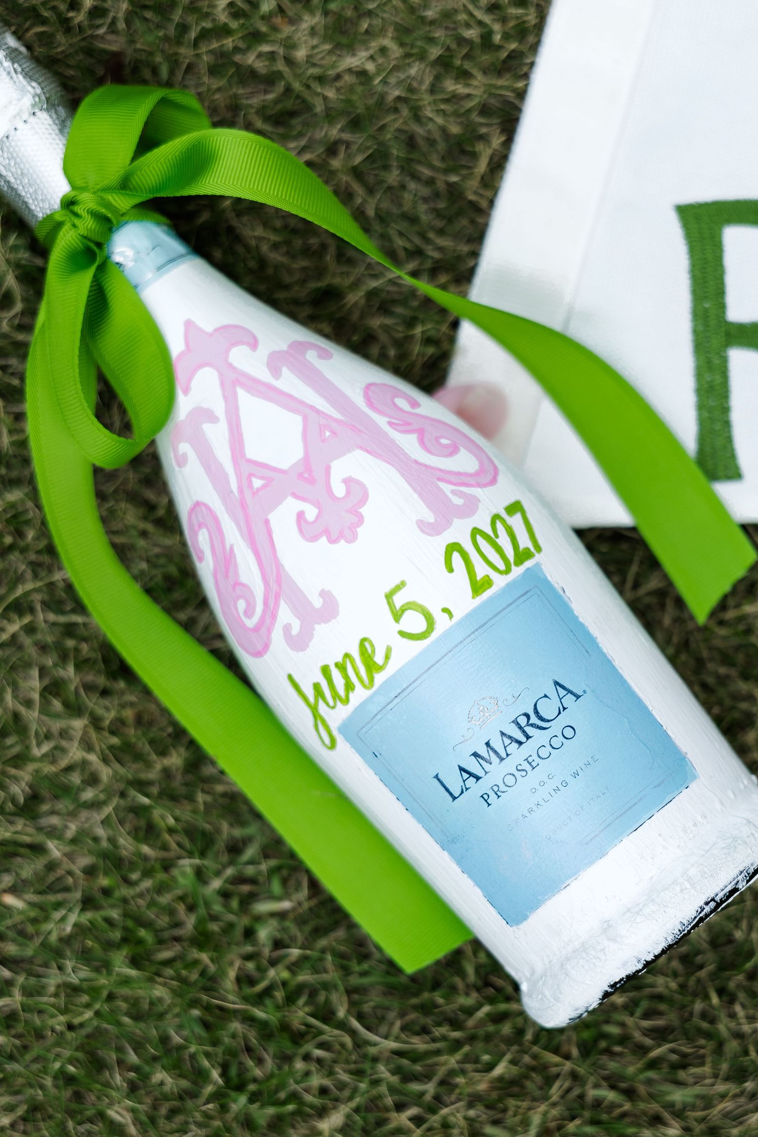 La Marca Prosecco bottle with pink monogram, green ribbon, and June 5, 2027 date, lying on grass.
