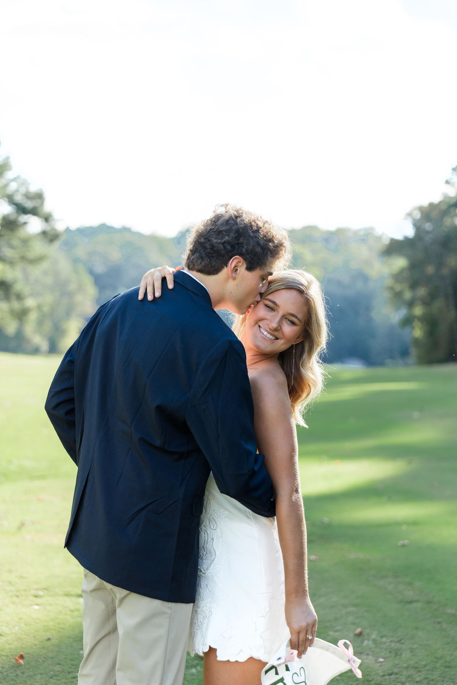 A smiling bride in a white dress embraces a groom in a navy blazer outdoors on a sunny green lawn.