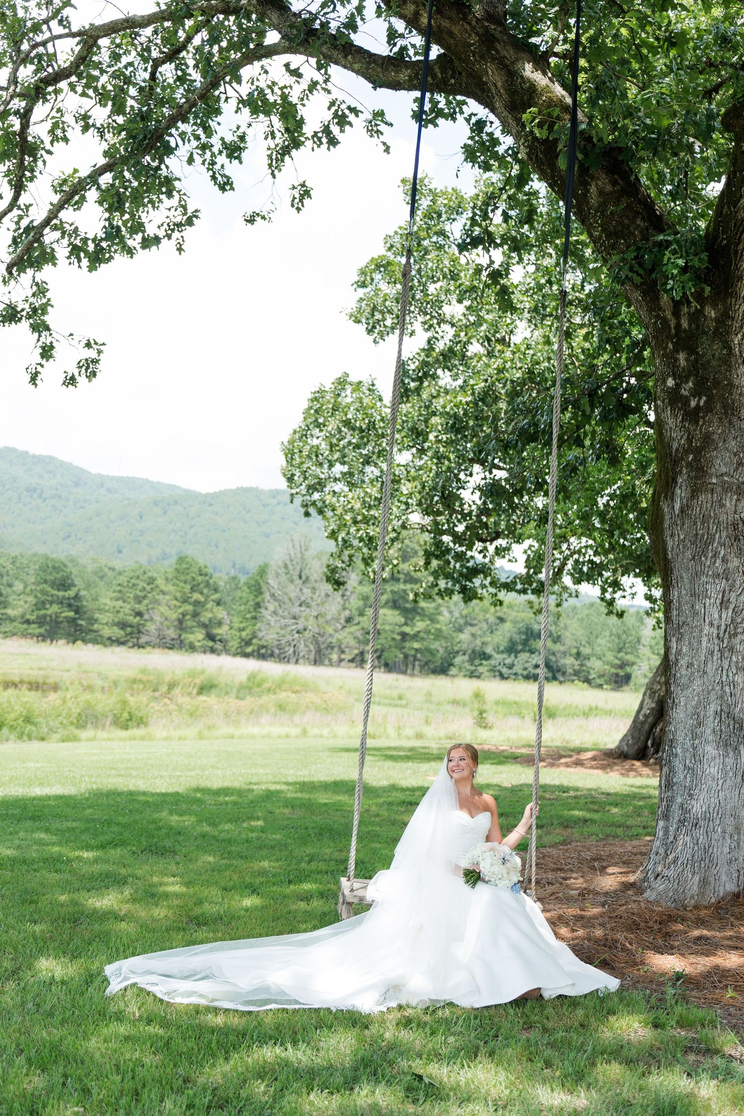 Bride in white gown sitting on a tree swing holding a bouquet, with scenic green hills in the background.