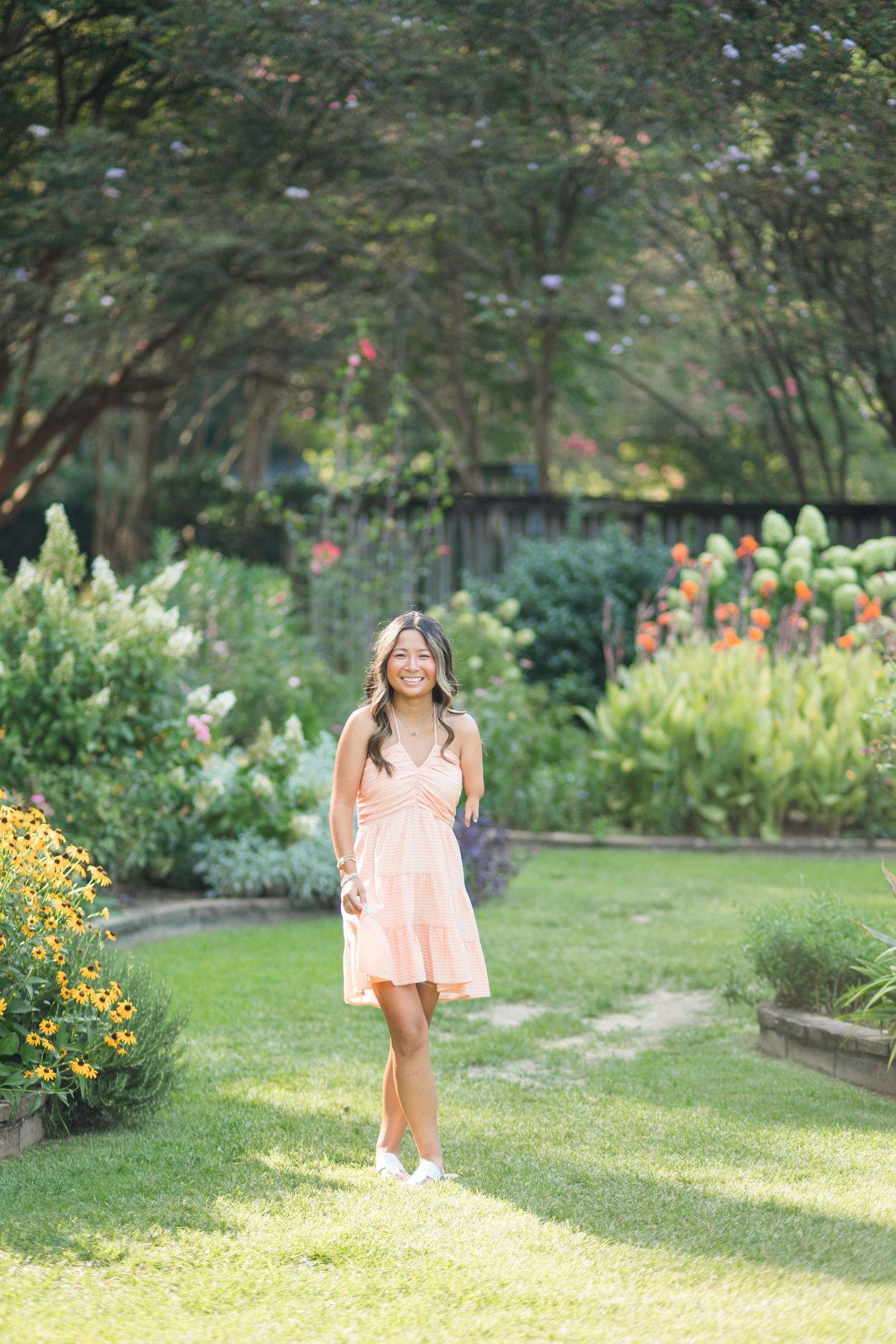 Young woman in a peach dress smiling in a lush garden with colorful flowers and green trees.