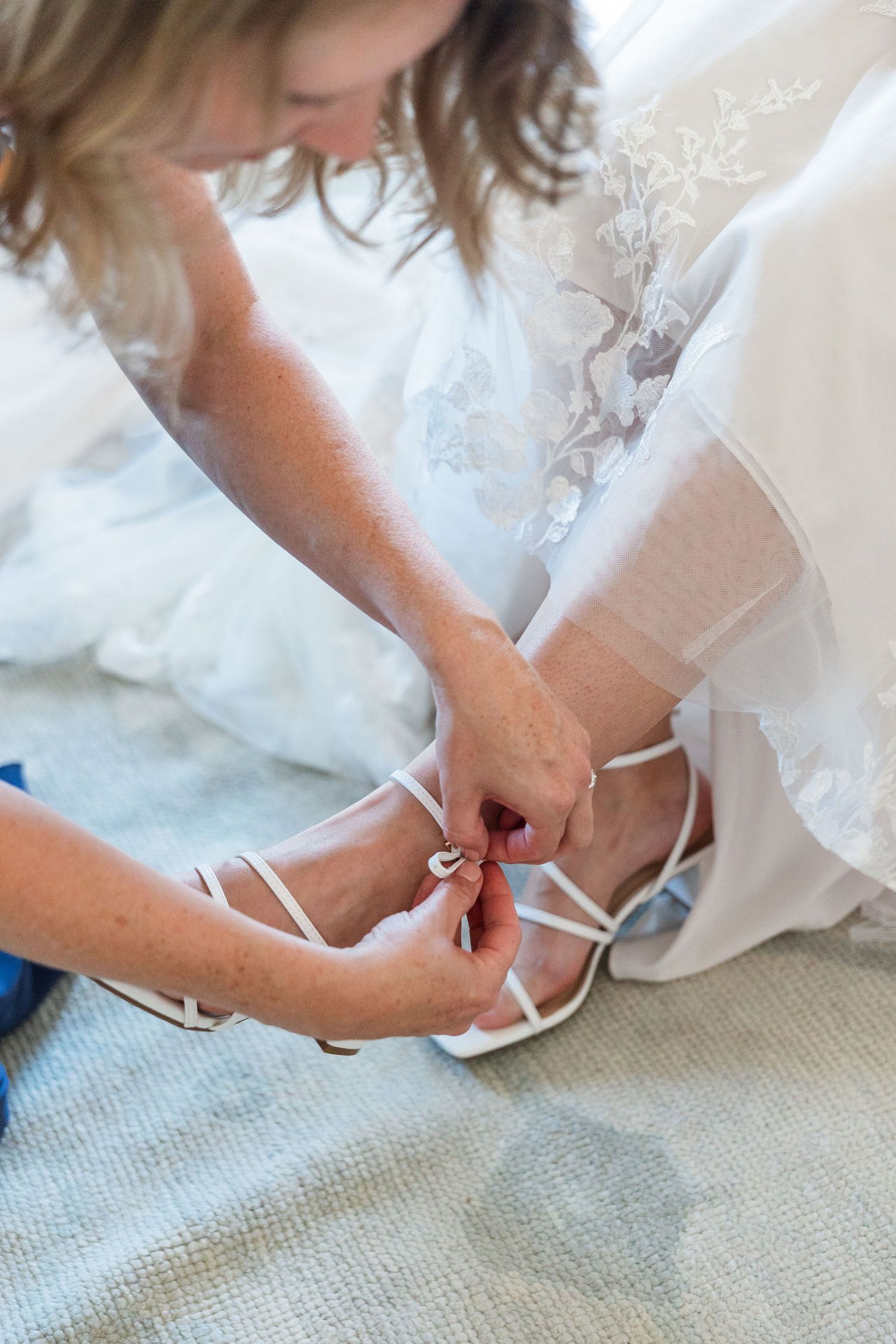 Bridesmaid helping bride fasten white strappy wedding heels before the ceremony.