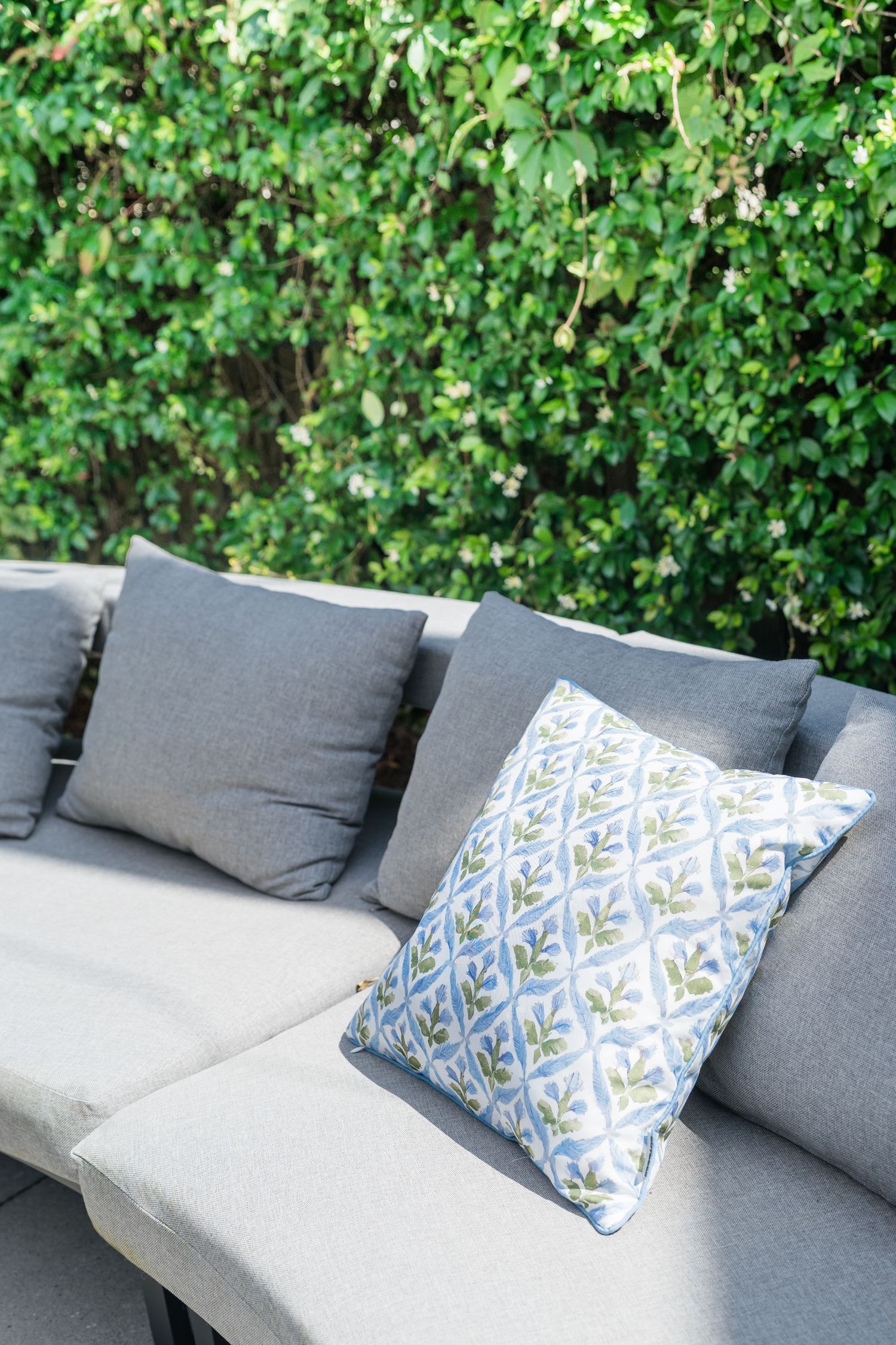 Gray outdoor patio sofa with decorative patterned pillow against lush green hedge wall.