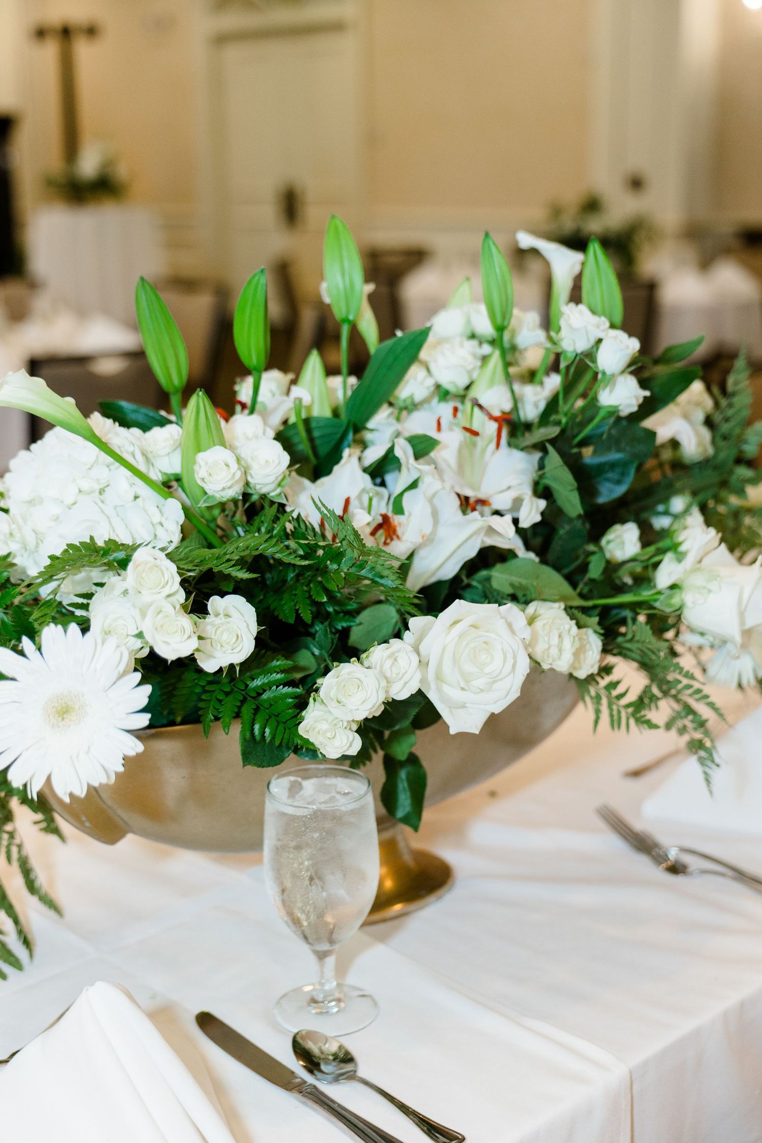 Lush white wedding centerpiece with roses, lilies, and greenery in a gold compote vase on a reception table.