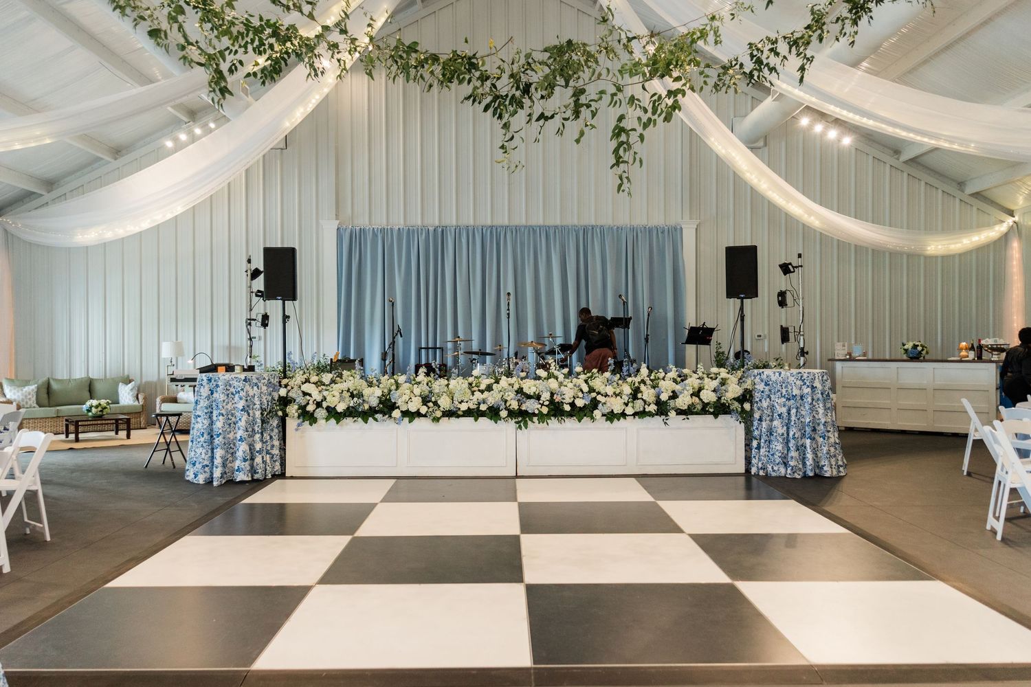 Wedding reception venue with black and white checkered dance floor, floral head table, and draped white ceiling decor.